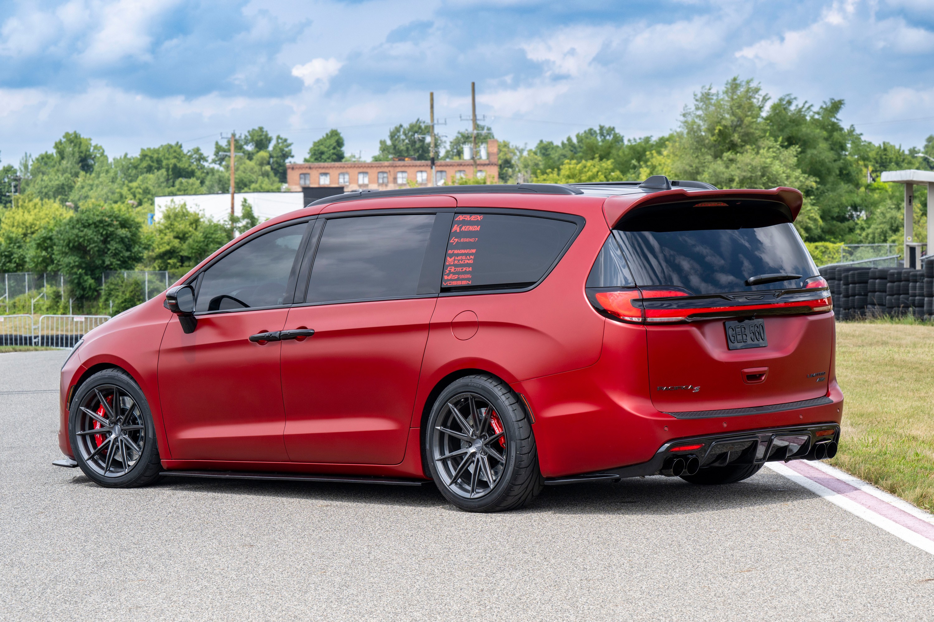 Minivan vermelha fosca, com rodas esportivas pretas e pinças de freio vermelhas, estacionada em uma pista de corrida. O veículo tem vidros escuros e adesivos na janela traseira. Ao fundo, árvores, um prédio de tijolos e um céu azul com nuvens brancas