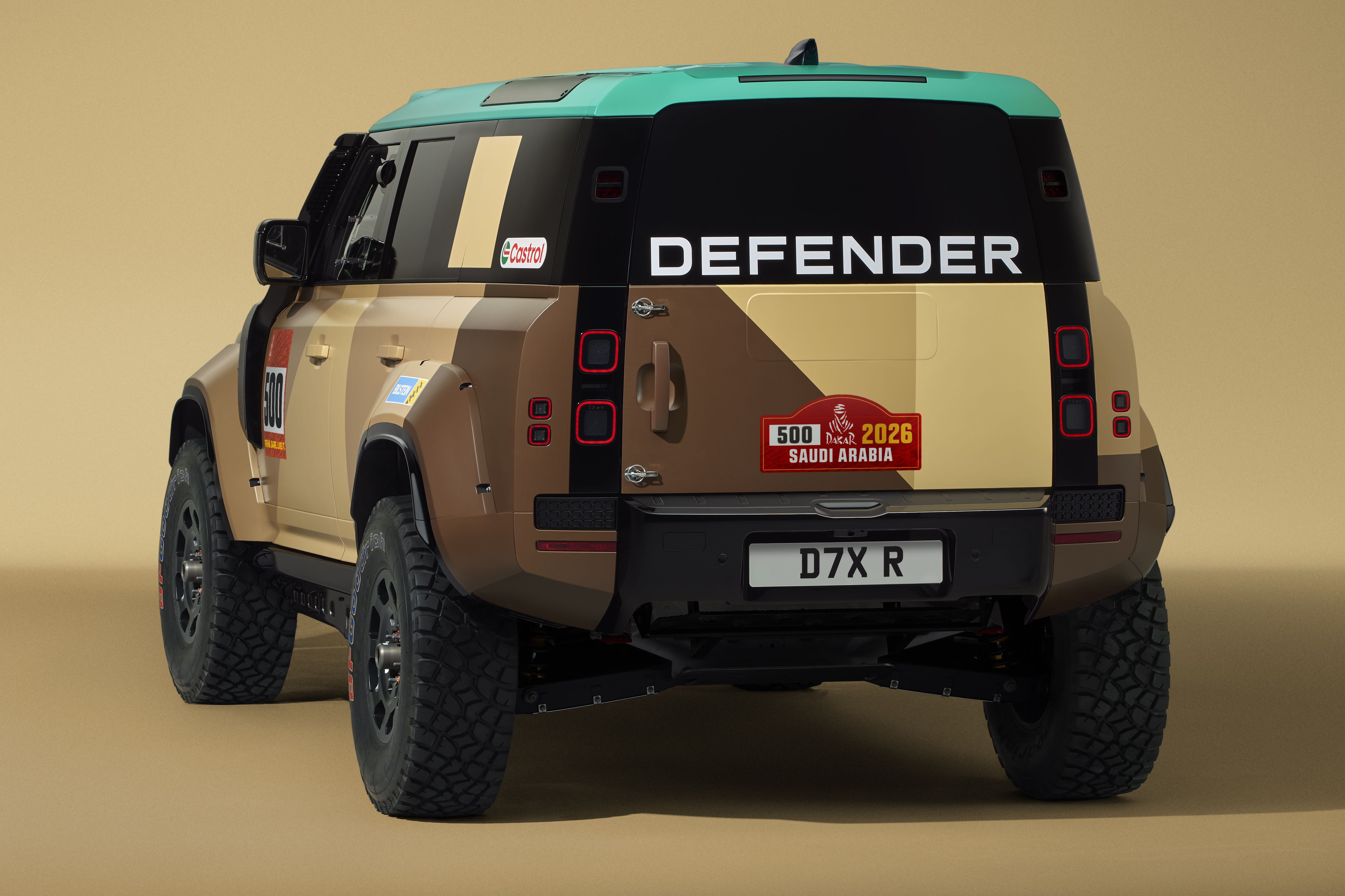 Traseira de um Land Rover Defender com pintura camuflada em tons de bege e marrom, teto verde-água. O nome DEFENDER está em branco na parte superior da porta traseira. Há um adesivo vermelho com 500 2026 SAUDI ARABIA e a placa D7X R na parte inferior. O veículo possui pneus robustos para off-road