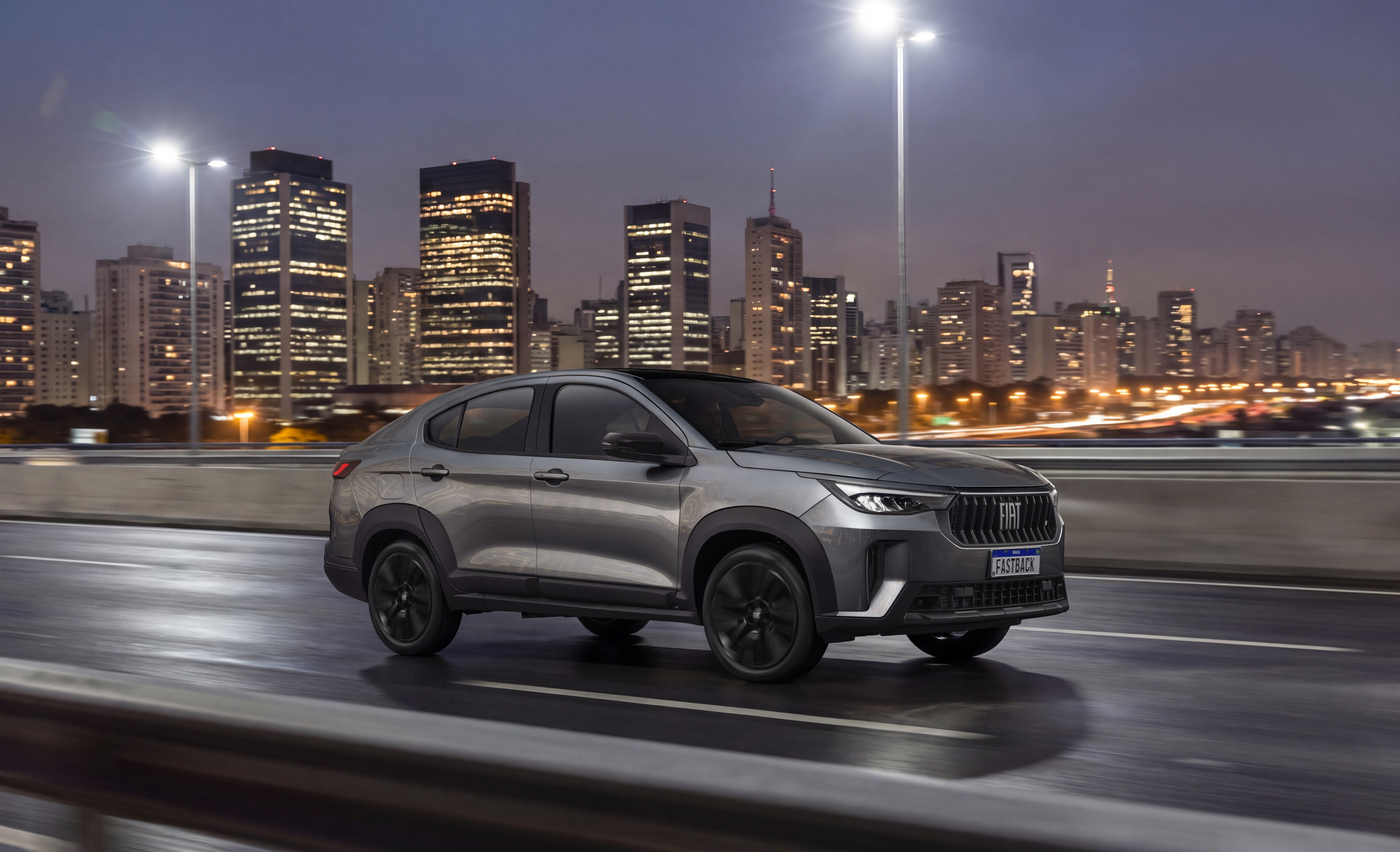 SUV cupê Fiat Fastback cinza escuro em movimento, à noite, com faróis acesos, em uma via expressa. Ao fundo, prédios iluminados de uma cidade grande e postes de luz.