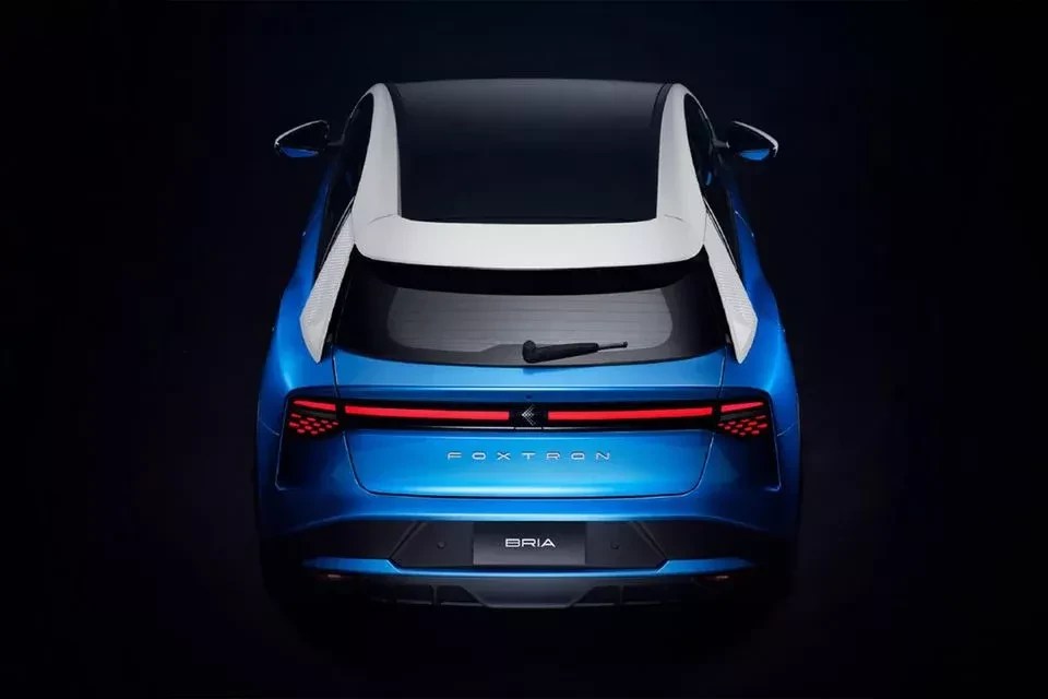 Traseira de um carro elétrico azul-metálico, modelo Foxtron Bria, com teto preto e detalhes brancos nas laterais. As lanternas traseiras são uma faixa horizontal vermelha que se estende por toda a largura do veículo, com padrões de pontos luminosos nas extremidades. O limpador de para-brisa está na parte superior do vidro traseiro. O carro está em um fundo escuro, com iluminação focada na parte traseira