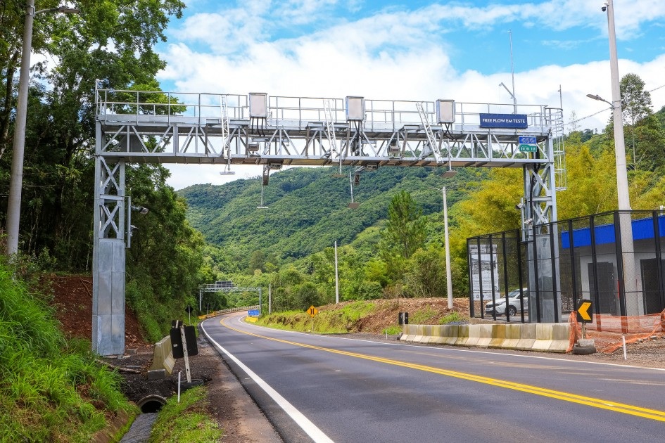 Pórtico de pedágio free flow sobre uma rodovia sinuosa, com montanhas verdes ao fundo e céu azul com nuvens. À direita, uma estrutura azul e preta cercada por grades.