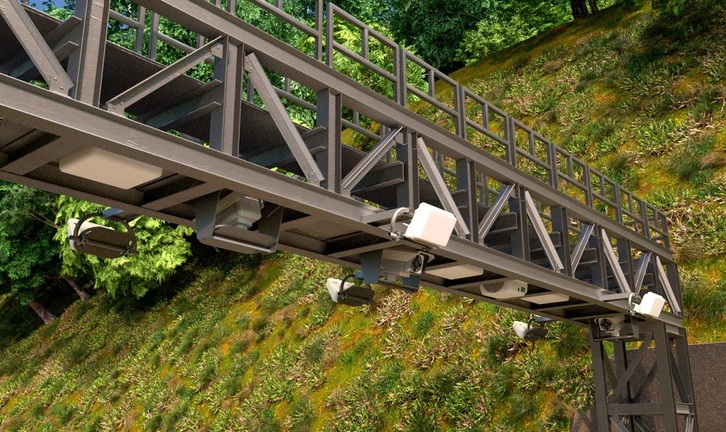 Estrutura metálica de uma passarela ou pórtico de pedágio eletrônico, com câmeras e sensores instalados na parte inferior, sobre uma estrada escura. Ao fundo, um morro coberto por vegetação verde e árvores