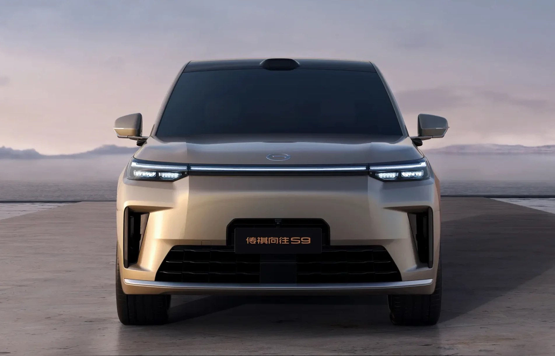 Carro dourado, modelo SUV, visto de frente, com faróis em LED acesos e uma barra de luz horizontal. Ao fundo, um cenário de montanhas e céu claro, com estruturas retangulares de vidro nas laterais
