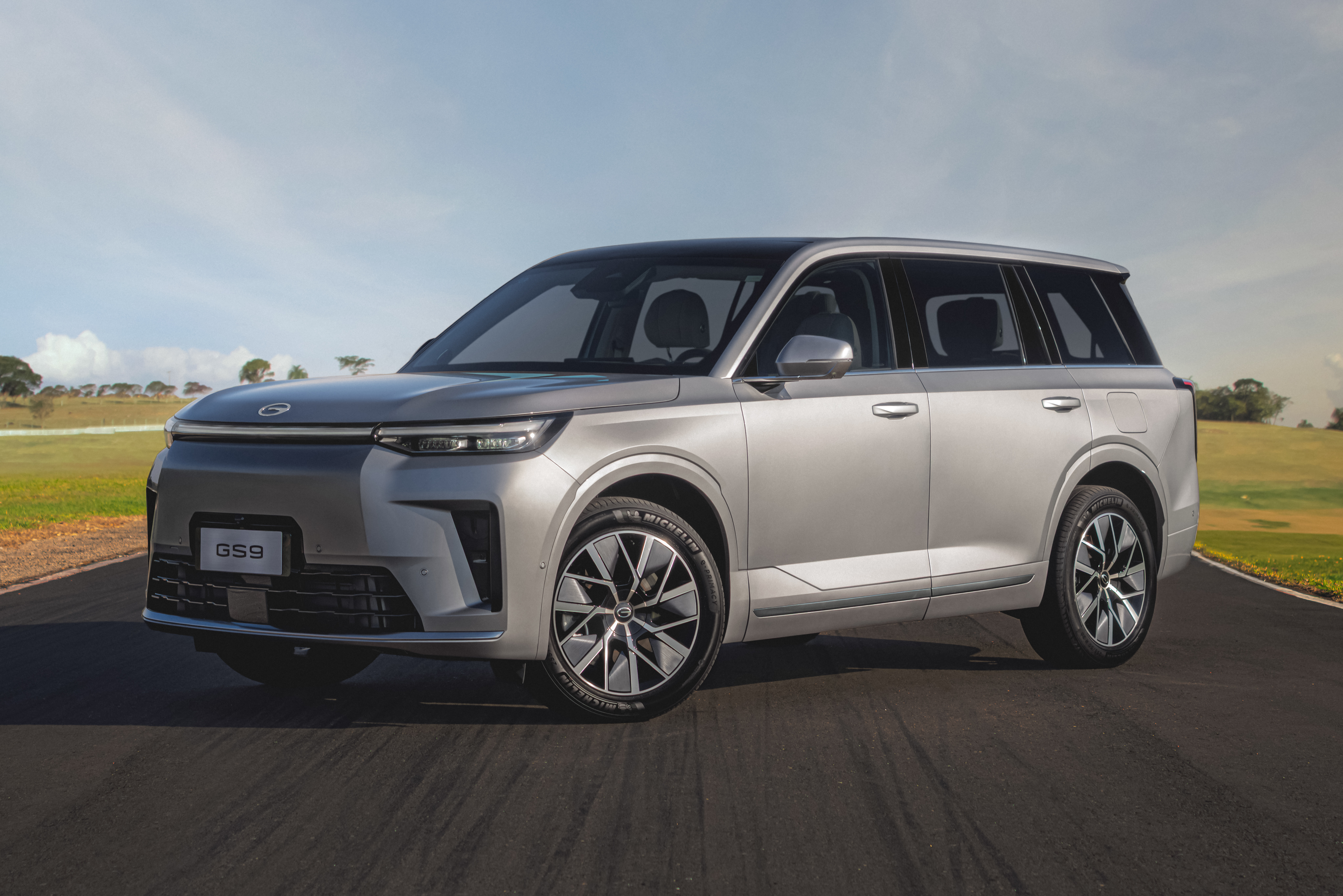 SUV GAC GS9 prateado, com teto preto, estacionado em uma estrada asfaltada. Ao fundo, um campo verde e c&eacute;u azul com nuvens esparsas
