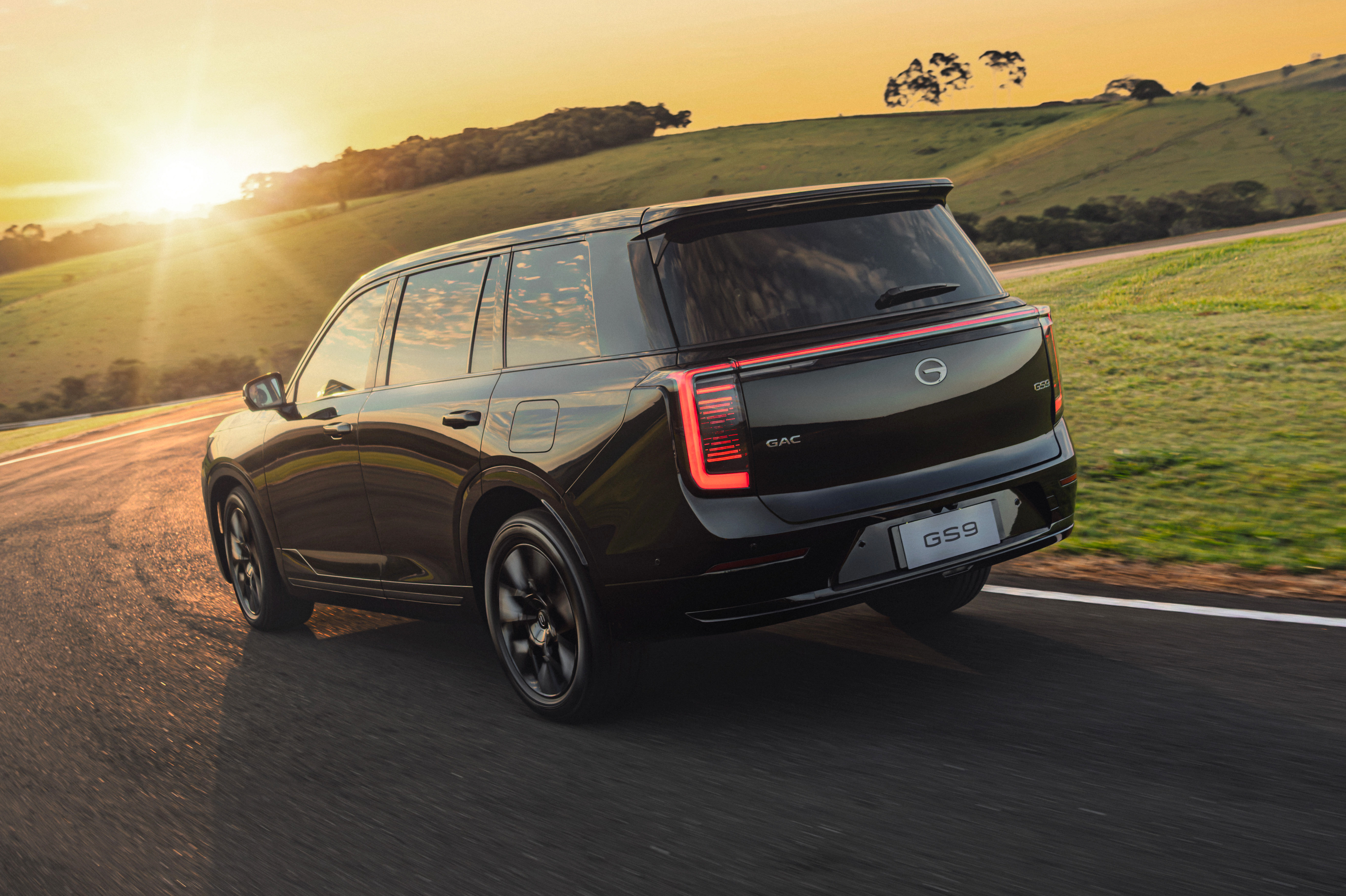 SUV preto GAC GS9 em uma estrada sinuosa, visto de tr&aacute;s, com as luzes traseiras acesas. O sol se p&otilde;e no horizonte, iluminando o c&eacute;u laranja e as colinas verdes ao fundo