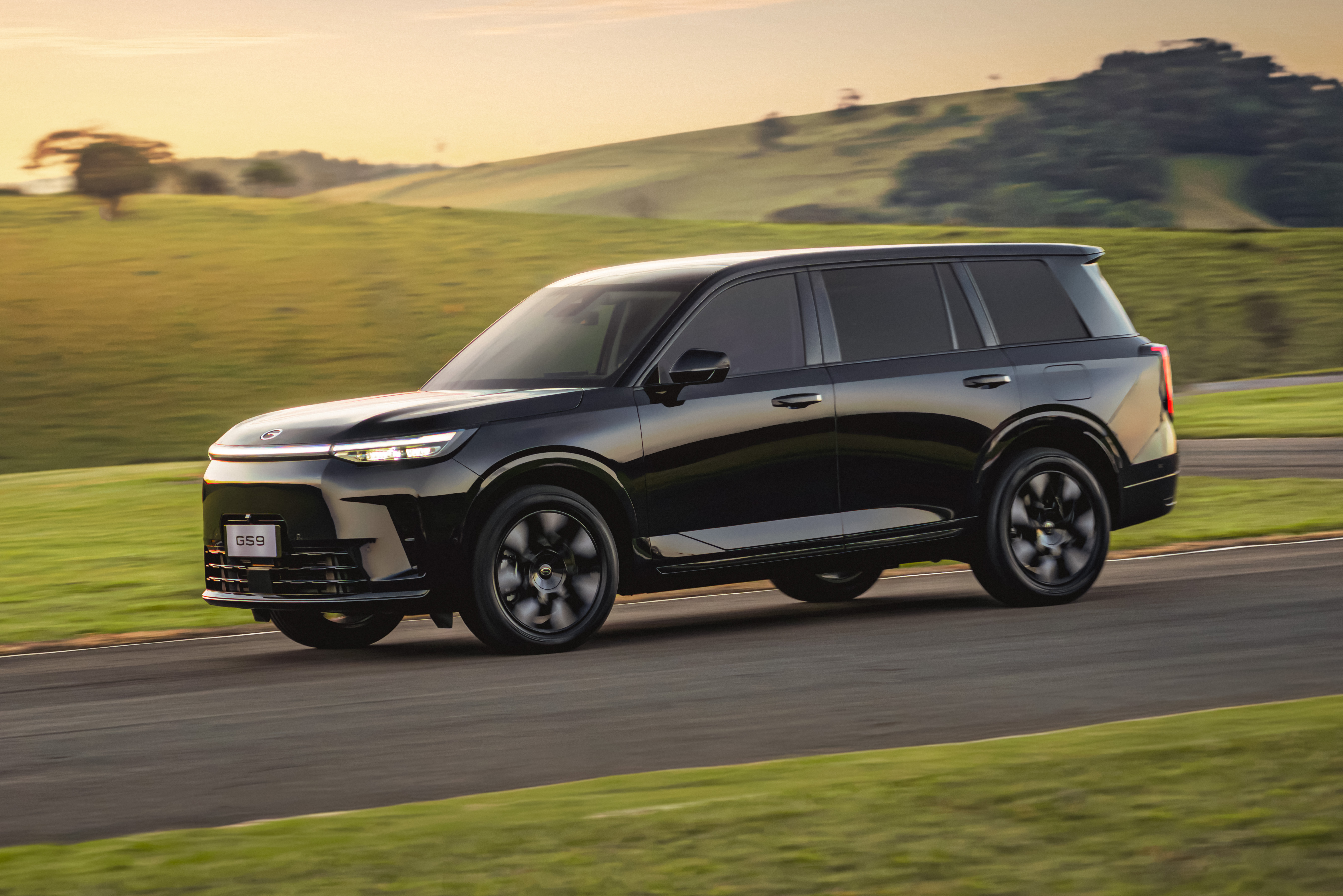 SUV preto brilhante em movimento, visto de lado, com rodas escuras e vidros fum&ecirc;. O ve&iacute;culo est&aacute; em uma estrada asfaltada, ladeada por gramados verdes e colinas ao fundo, sob um c&eacute;u alaranjado de p&ocirc;r do sol