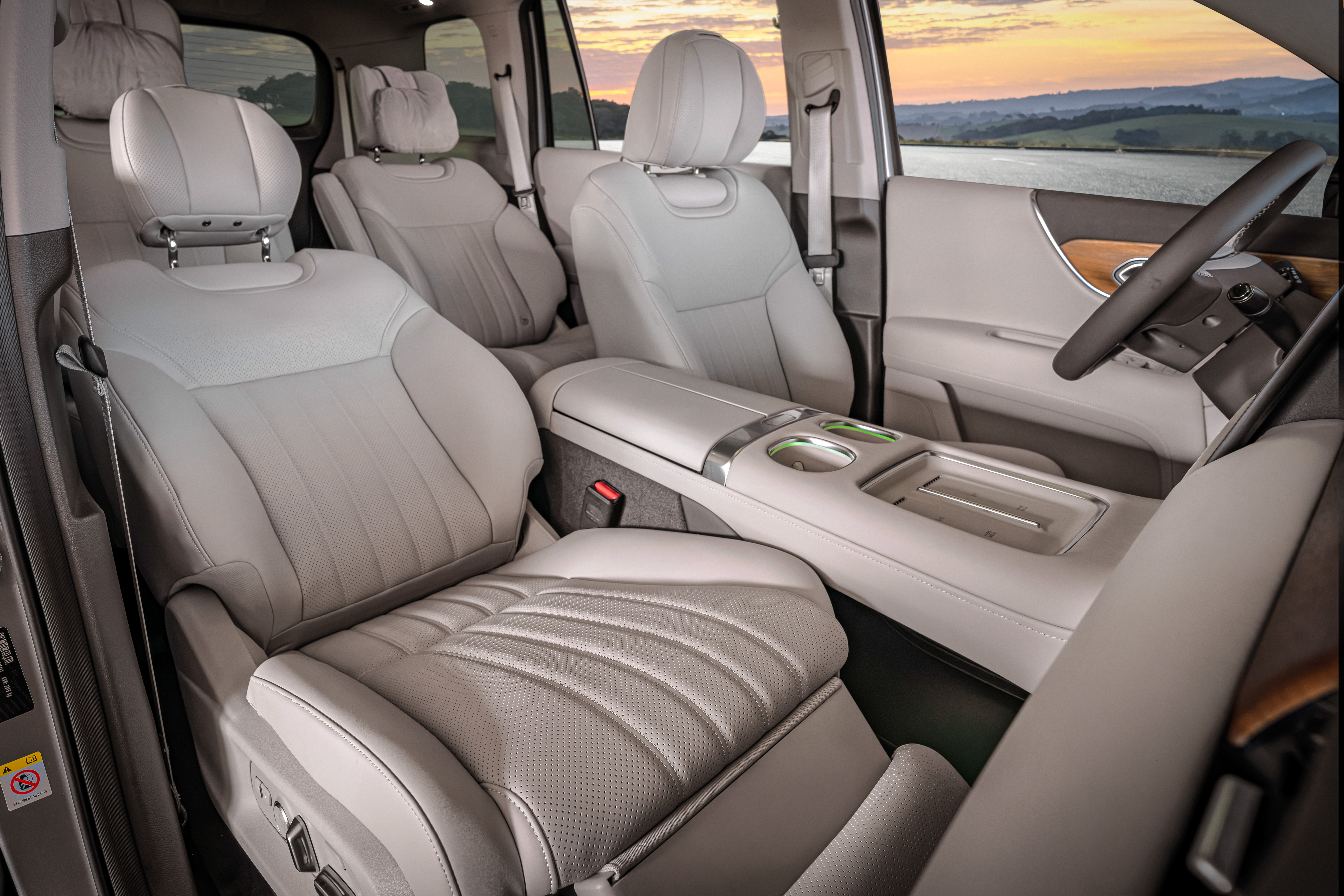 Interior de um carro de luxo com bancos de couro cinza-claro, console central com porta-copos e compartimento. Ao fundo, volante escuro e paisagem com lago e montanhas sob c&eacute;u alaranjado