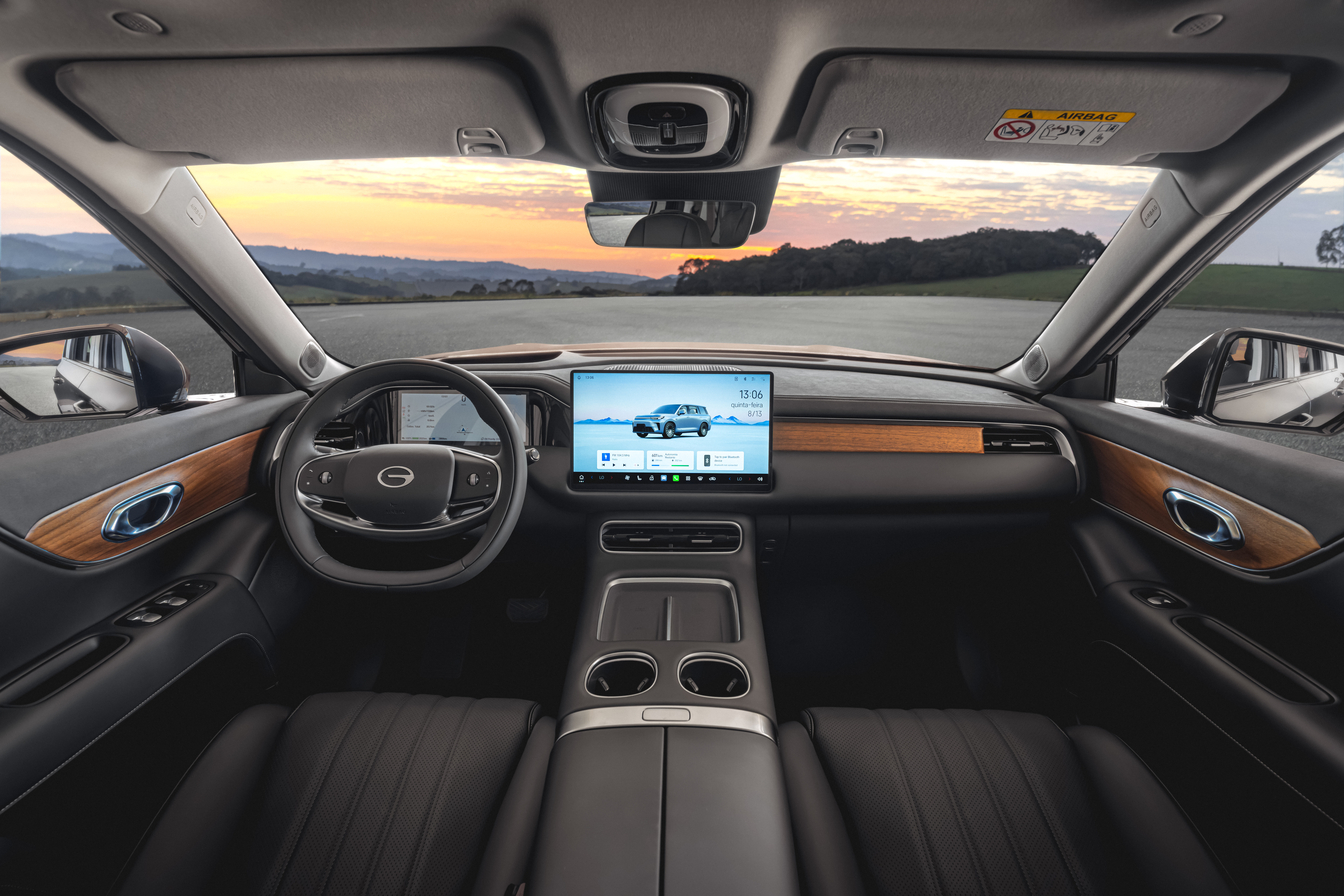 Interior de um carro moderno, com painel escuro e detalhes em madeira. O volante tem o logo da GAC. Uma tela central exibe um SUV cinza e informa&ccedil;&otilde;es do ve&iacute;culo. O para-brisa mostra um p&ocirc;r do sol sobre montanhas e uma estrada. Os bancos s&atilde;o de couro preto.