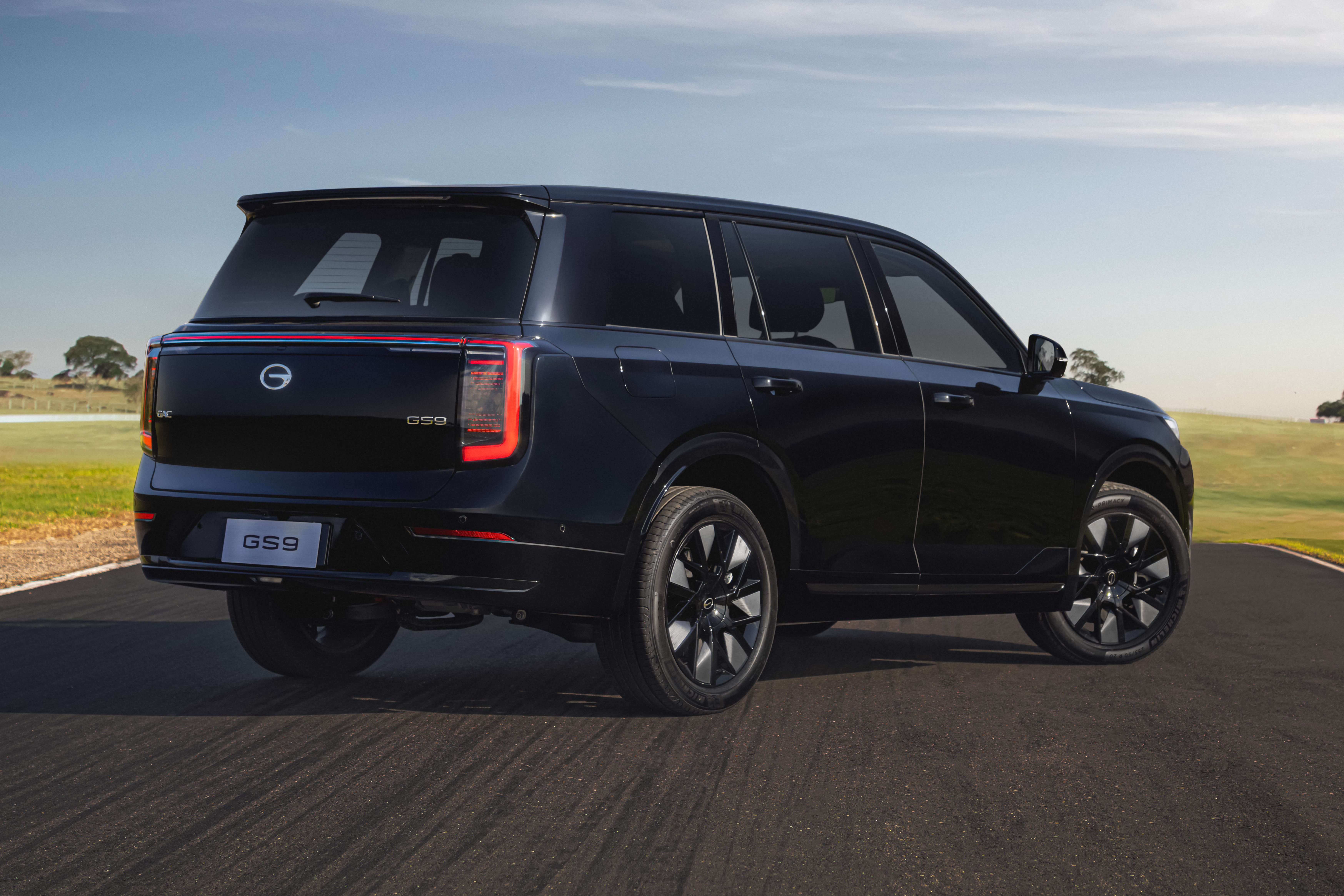 SUV GAC GS9 preto, visto de traseira e lateral, parado em uma estrada asfaltada com gramado verde e c&eacute;u azul ao fundo