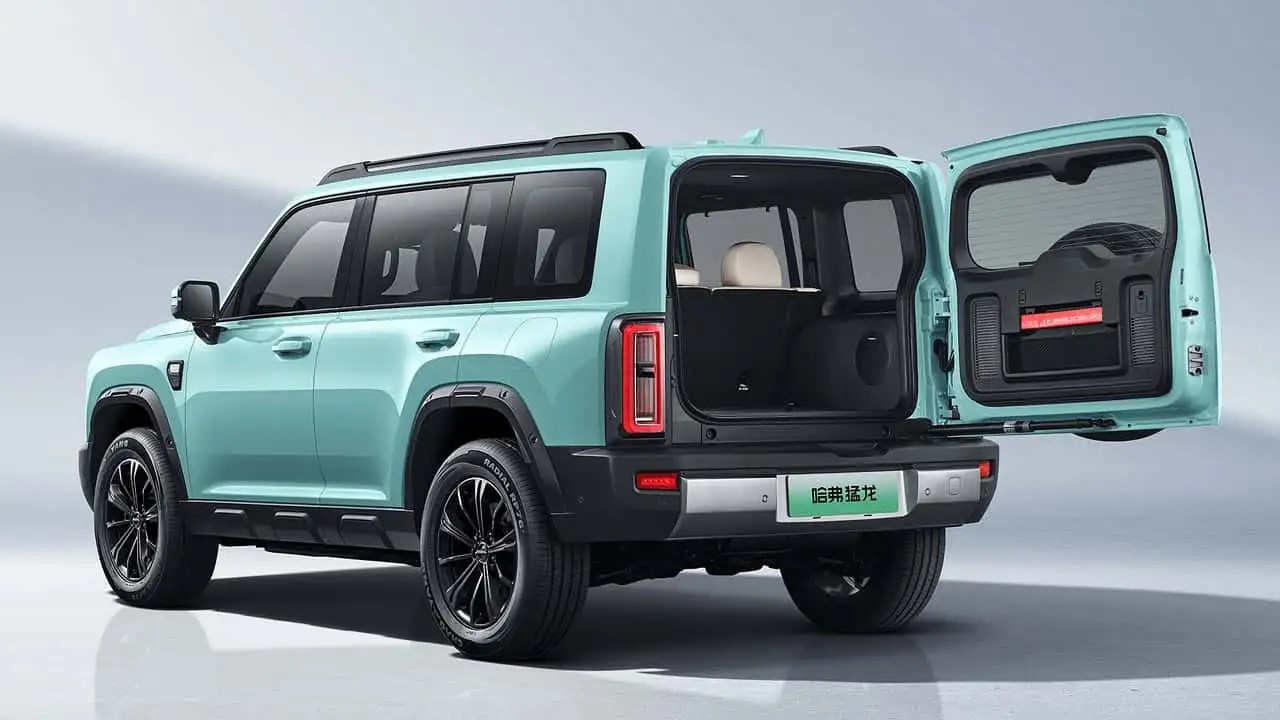 SUV verde-água com porta-malas aberto, revelando o interior espaçoso e o estepe na lateral. O veículo tem rodas pretas e detalhes robustos, em um fundo claro