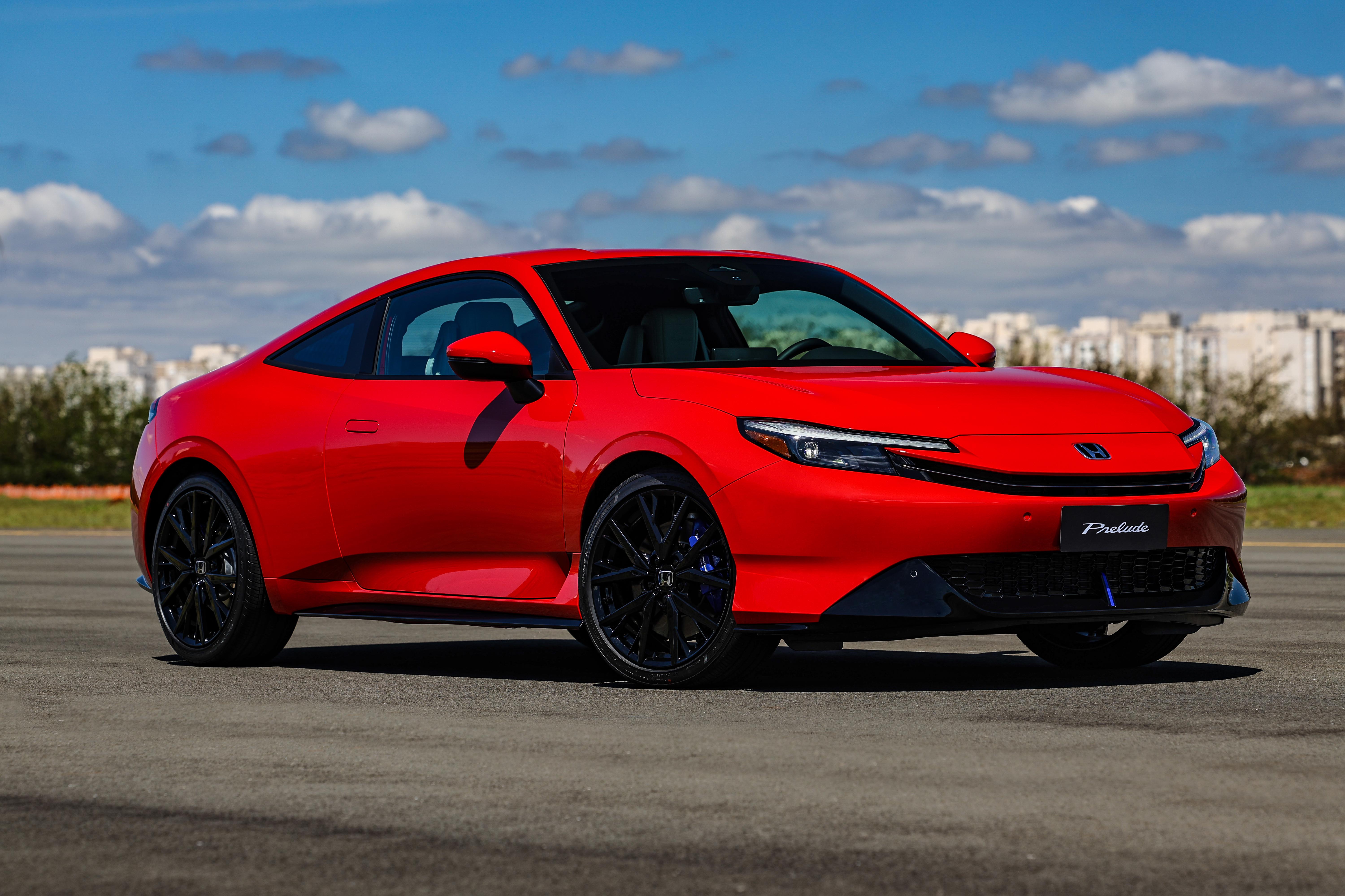Carro esportivo vermelho brilhante, modelo Prelude, estacionado em uma pista de asfalto sob céu azul com nuvens esparsas. Ao fundo, prédios urbanos e vegetação.