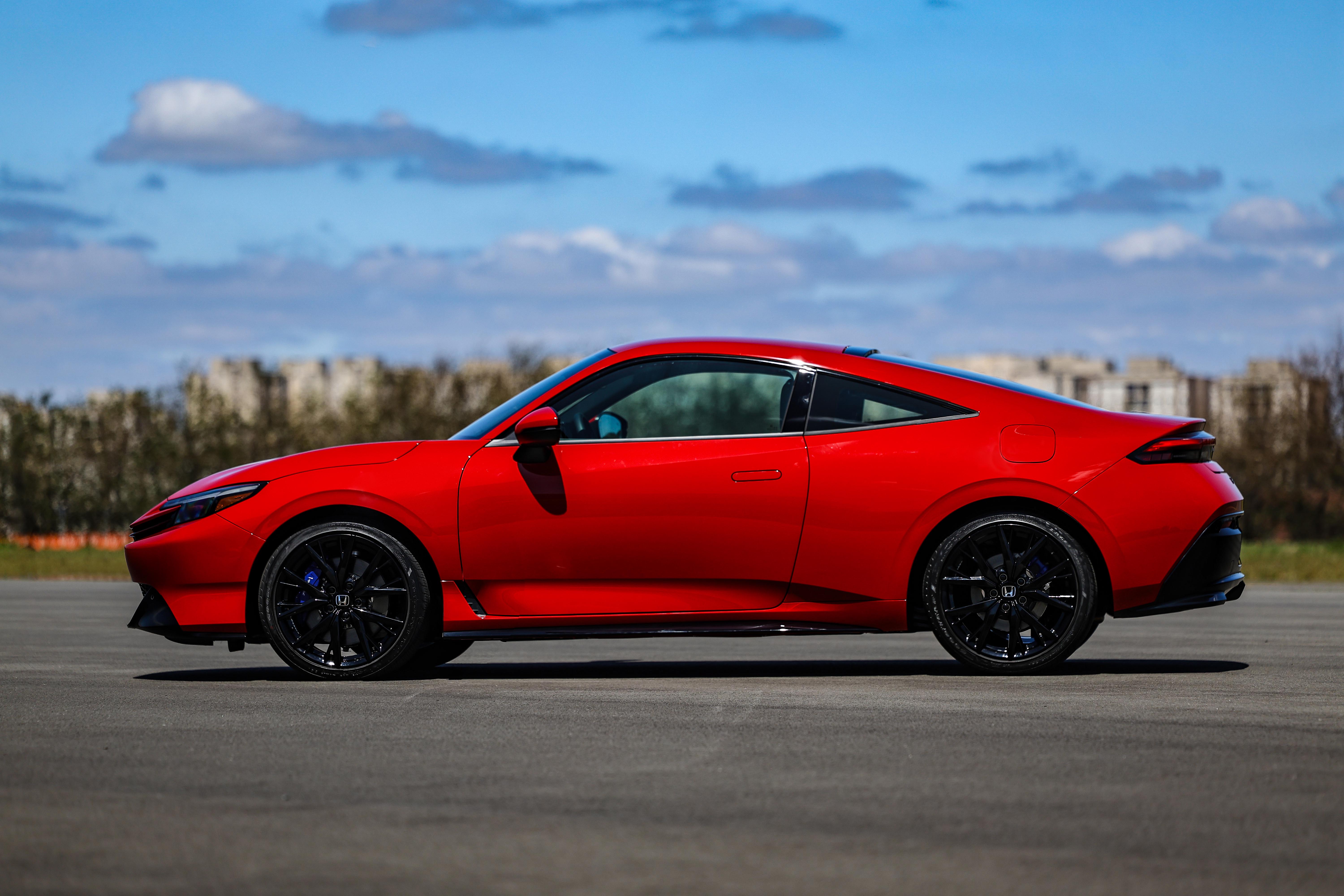 Carro esportivo vermelho brilhante, visto de lado, parado em uma pista de asfalto sob céu azul com nuvens esparsas. Rodas pretas com detalhes azuis nos freios.