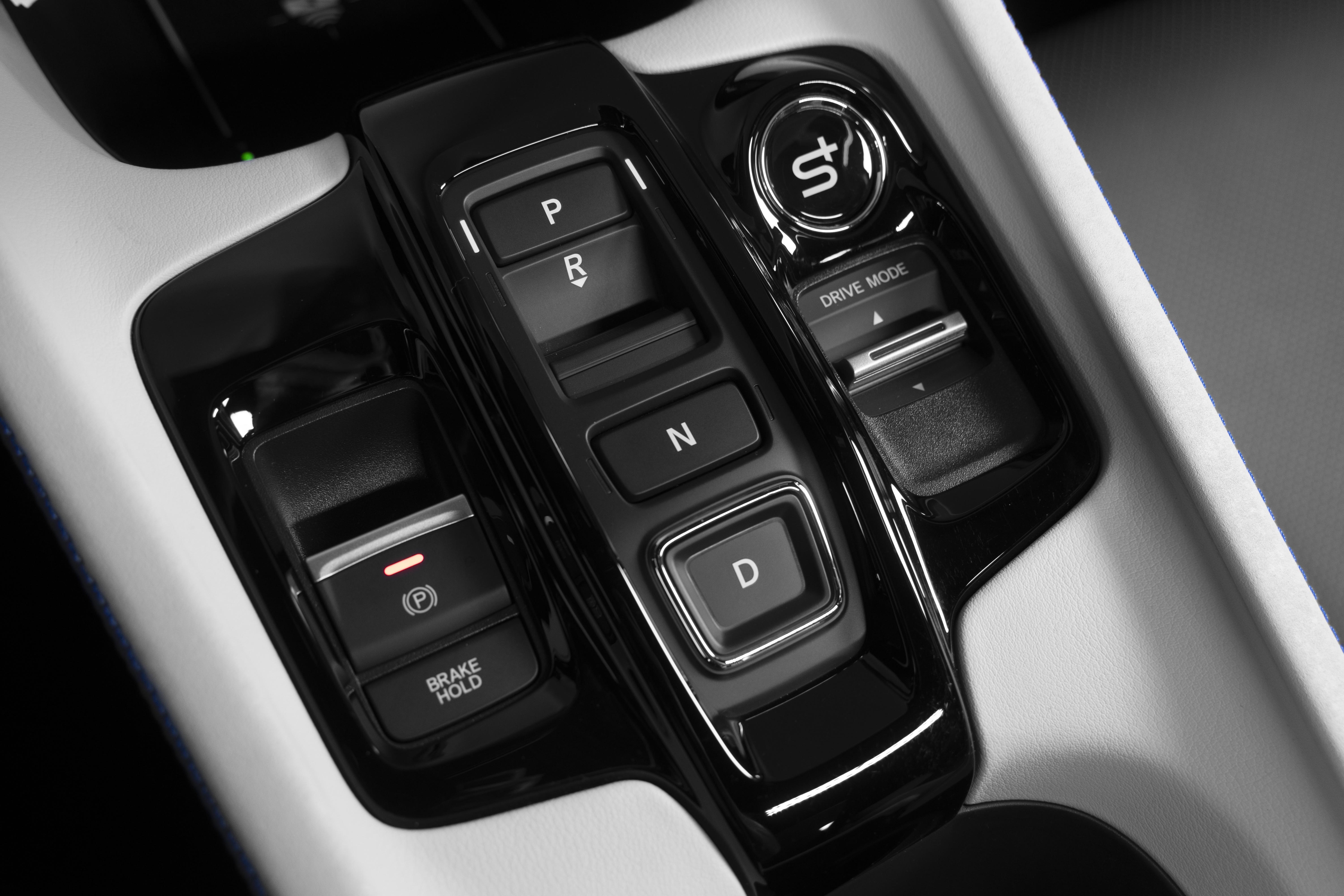 Painel de controle de um carro com botões pretos brilhantes em um console branco. Os botões incluem P, R, N, D, BRAKE HOLD, um botão de freio de estacionamento com luz vermelha, e um seletor DRIVE MODE