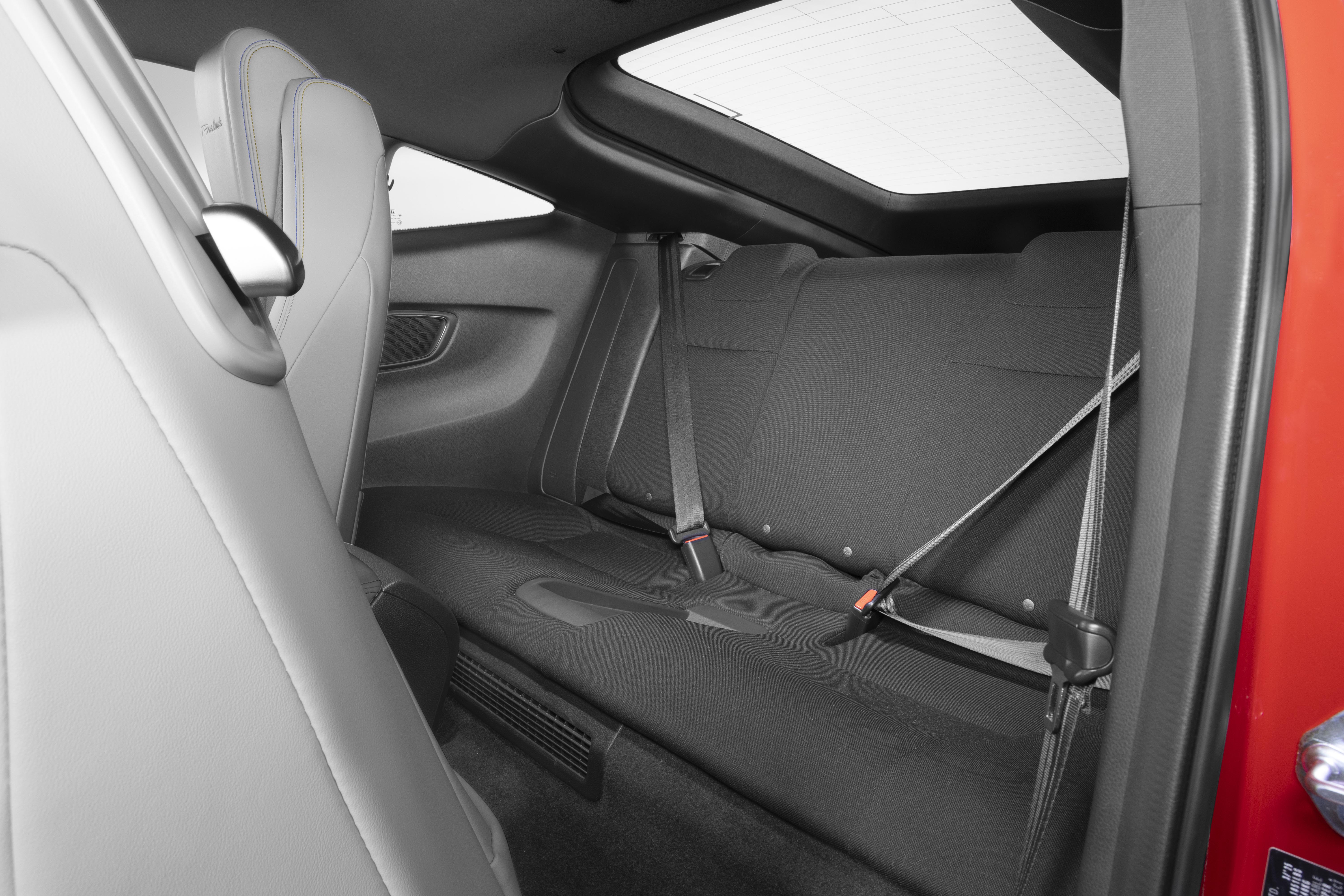 Interior de um carro esportivo com bancos traseiros pretos e cintos de segurança visíveis. O banco do passageiro dianteiro, em tom claro, aparece parcialmente à esquerda. Há um teto solar e uma janela traseira, com a porta vermelha do carro visível à direita