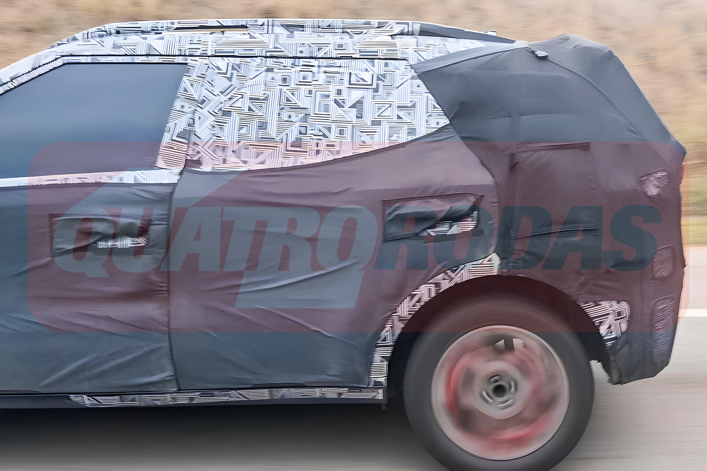 Lateral de um carro camuflado em movimento, com a parte superior coberta por adesivos geométricos brancos e pretos, e a parte inferior por tecido escuro. A roda traseira está borrada, indicando velocidade