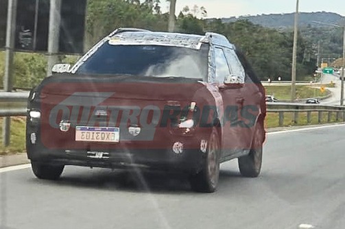 Carro protótipo camuflado em tons de vinho e preto, com teto branco, trafegando em uma rodovia. A placa dianteira está invertida, mostrando EX8I03. Ao fundo, uma paisagem montanhosa com vegetação densa e outras vias.