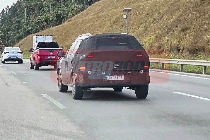 SUV camuflado em teste na estrada, visto por trás, com luzes traseiras acesas. Atrás dele, uma picape vermelha e um carro branco. À direita, um guard-rail e vegetação.