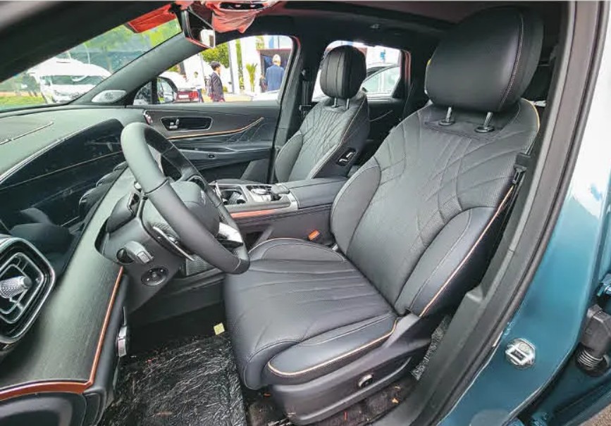 Interior de um carro moderno com bancos de couro preto e costura em losango, volante multifuncional e painel escuro. O banco do motorista está em primeiro plano, com detalhes em costura clara e ajuste elétrico. Ao fundo, pessoas e um barco branco são visíveis através dos vidros