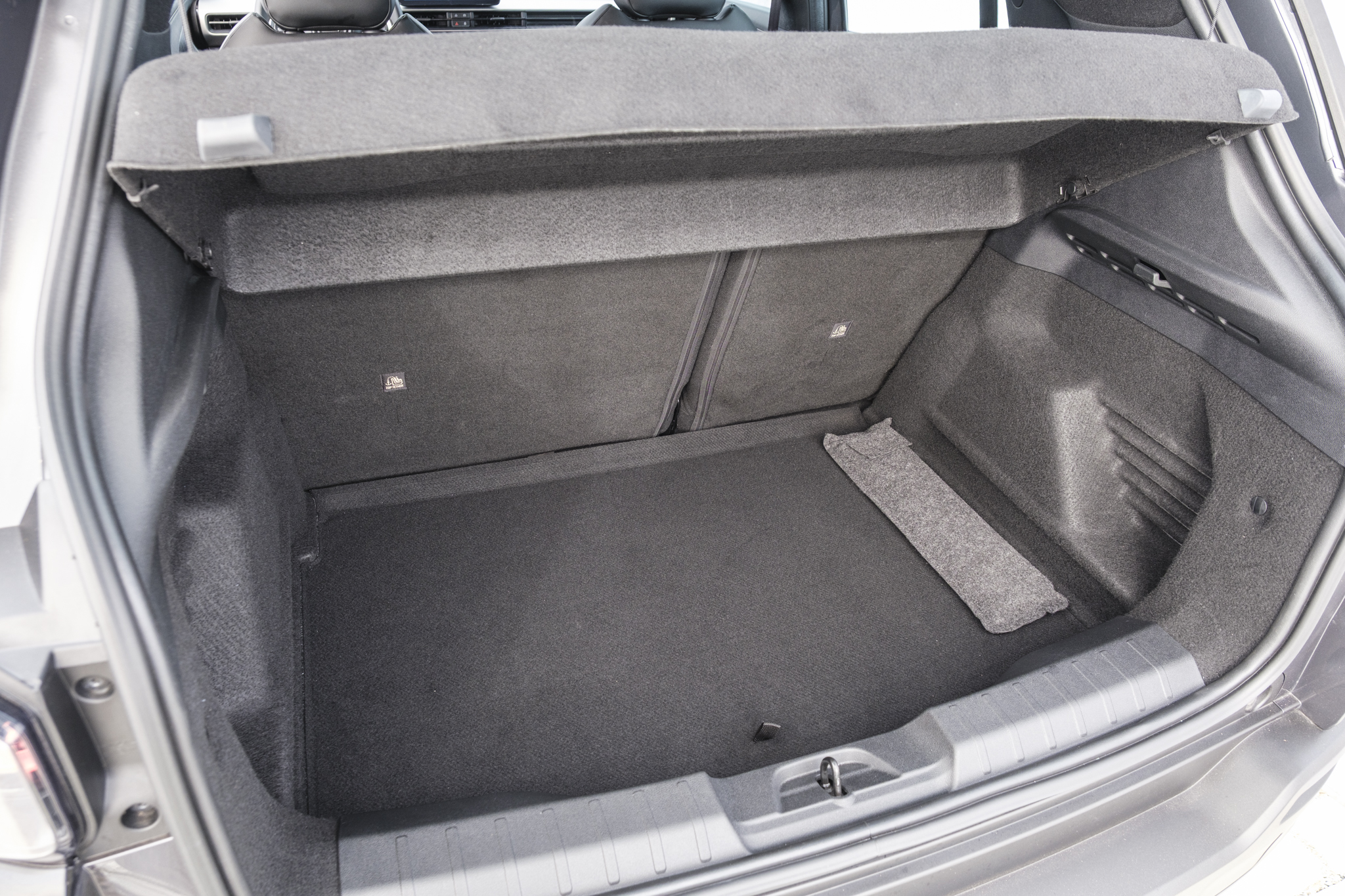 Porta-malas de carro cinza escuro, vazio e limpo, com o tampão superior levantado. O assoalho e as laterais são revestidos com carpete, e há um tapete retangular cinza claro no canto direito. O banco traseiro, também cinza, está visível ao fundo