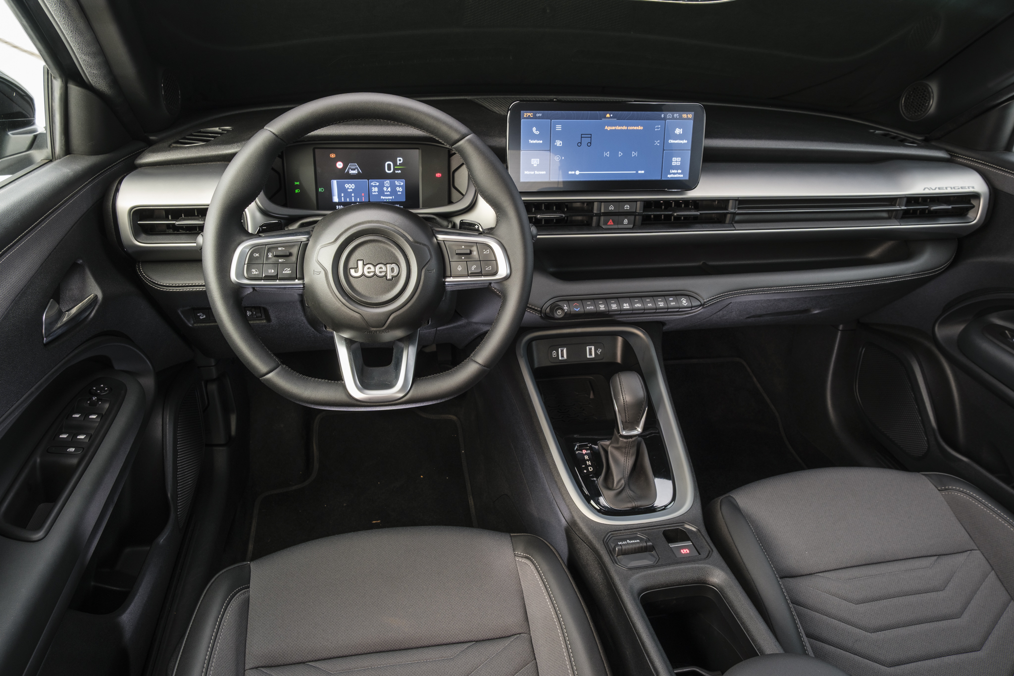 Interior de um carro moderno, com volante preto e o logo Jeep ao centro. Painel digital exibe informações do veículo, e uma tela multimídia sensível ao toque mostra um aplicativo de música. Bancos de couro escuro com detalhes em cinza, câmbio automático e console central com porta-objetos completam o ambiente. A porta do motorista tem controles dos vidros elétricos