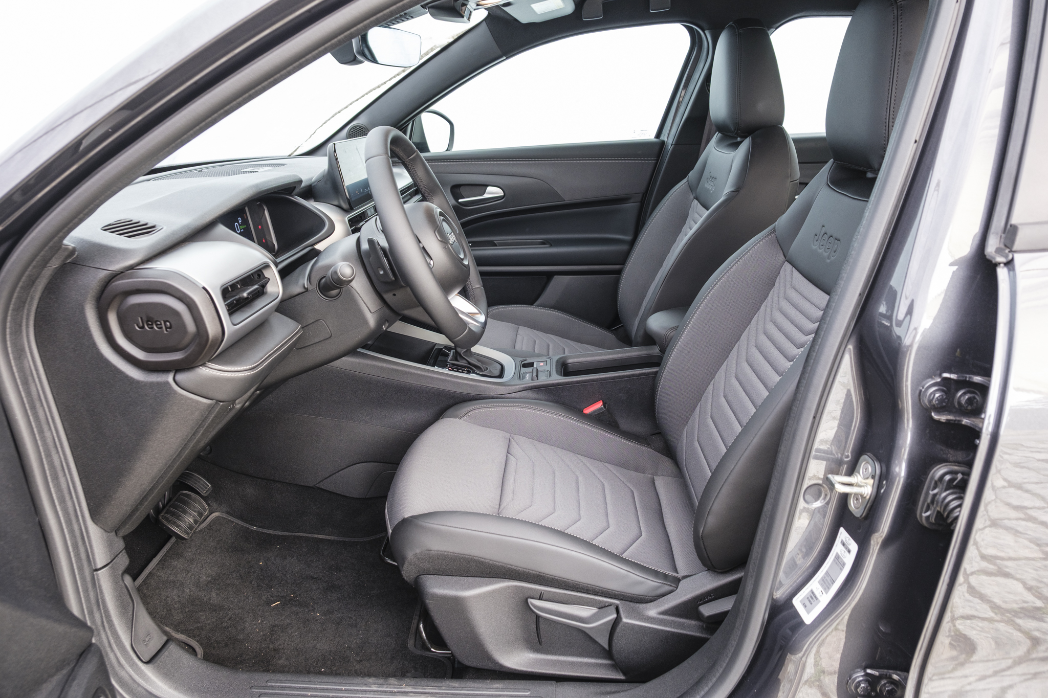 Interior de um carro Jeep, mostrando o banco do motorista e passageiro dianteiro em tons de cinza e preto, com detalhes texturizados. O volante escuro e o painel com saídas de ar e o logo Jeep são visíveis. O tapete preto e a porta aberta completam a cena