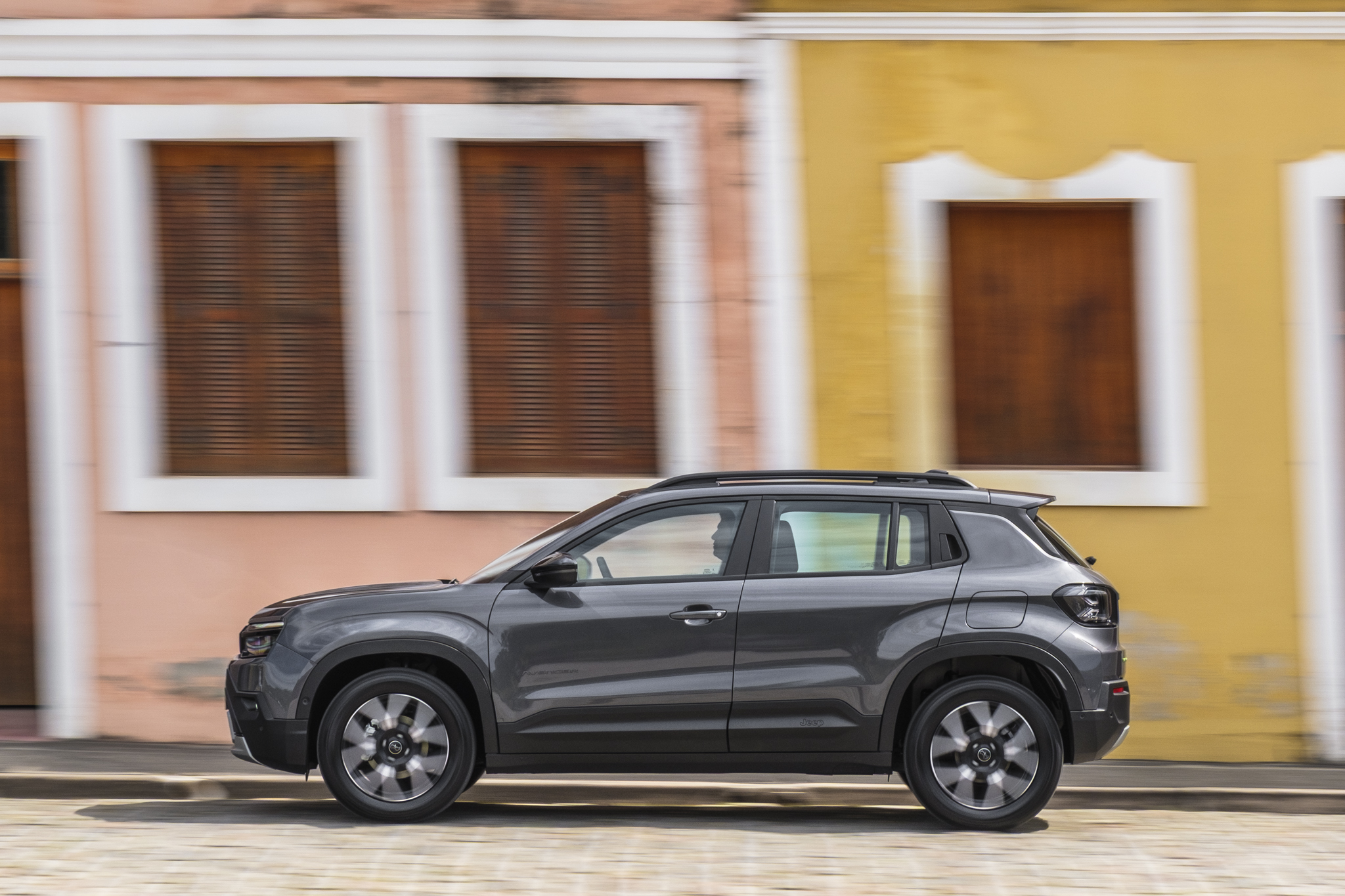 SUV cinza escuro em movimento, visto de lado, com um motorista visível. O fundo desfocado mostra fachadas de casas coloridas, uma rosa e outra amarela, com janelas de madeira escura e molduras brancas