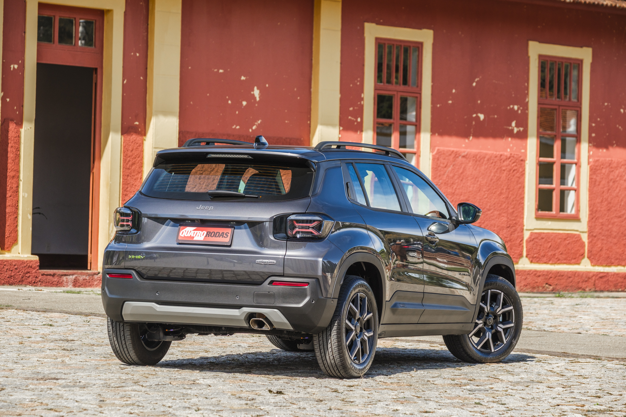 SUV cinza-escuro, visto de traseira e lateral, estacionado em rua de paralelepípedos. Ao fundo, uma parede vermelha com uma porta aberta e duas janelas de madeira marrom. O carro tem rack de teto e placa vermelha com Quatro Rodas