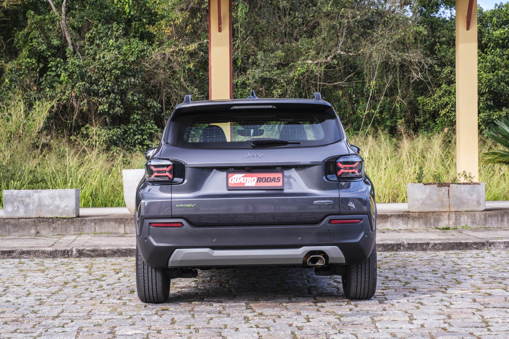 Traseira de um Jeep Avenger cinza escuro estacionado em piso de paralelepípedos, com a placa Quatro Rodas em vermelho. Ao fundo, vegetação densa e um pilar bege claro. As lanternas traseiras têm formato de X