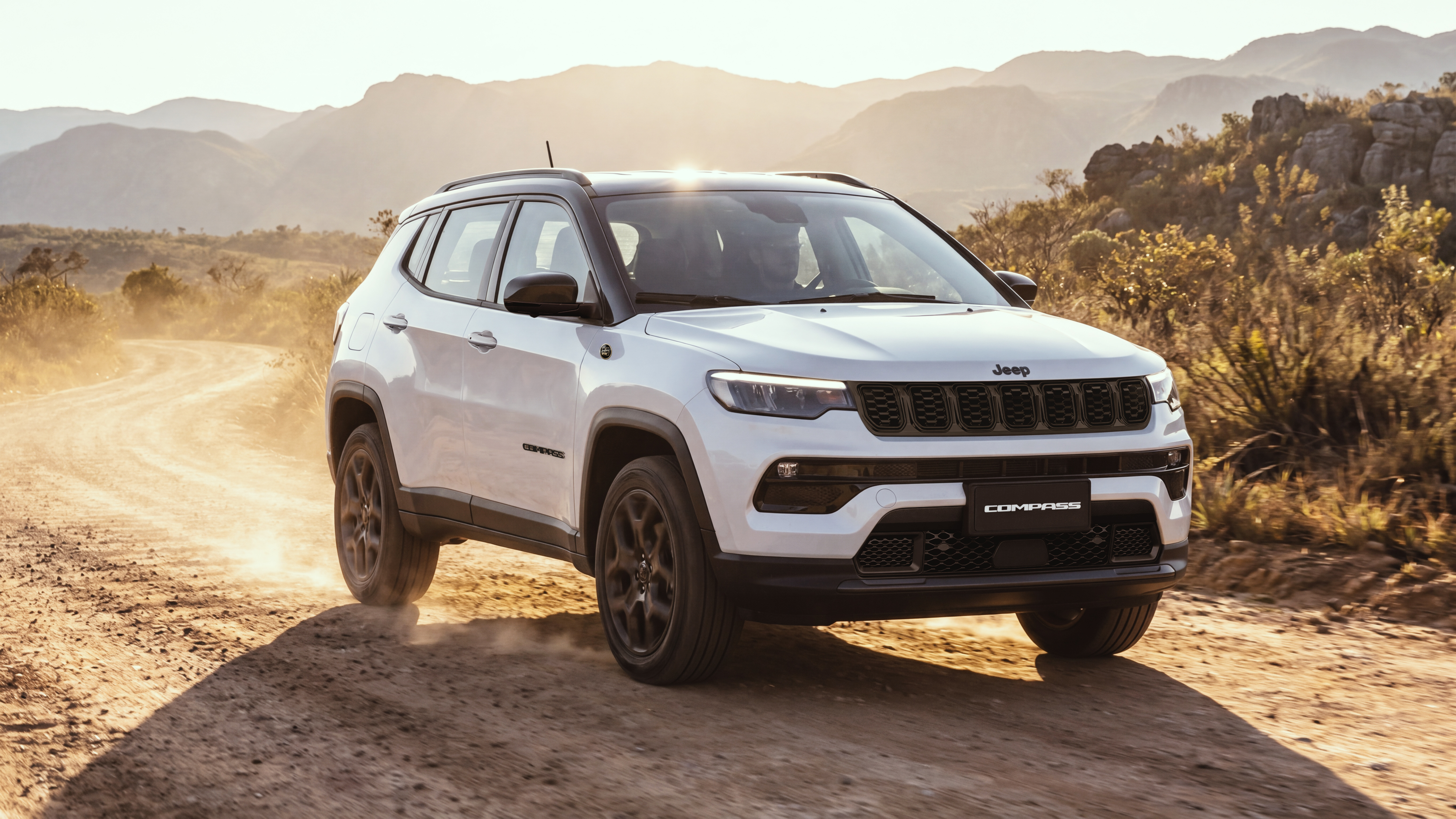 SUV Jeep Compass branco com teto preto, em estrada de terra com poeira, sob sol forte. Montanhas e vegetação seca ao fundo