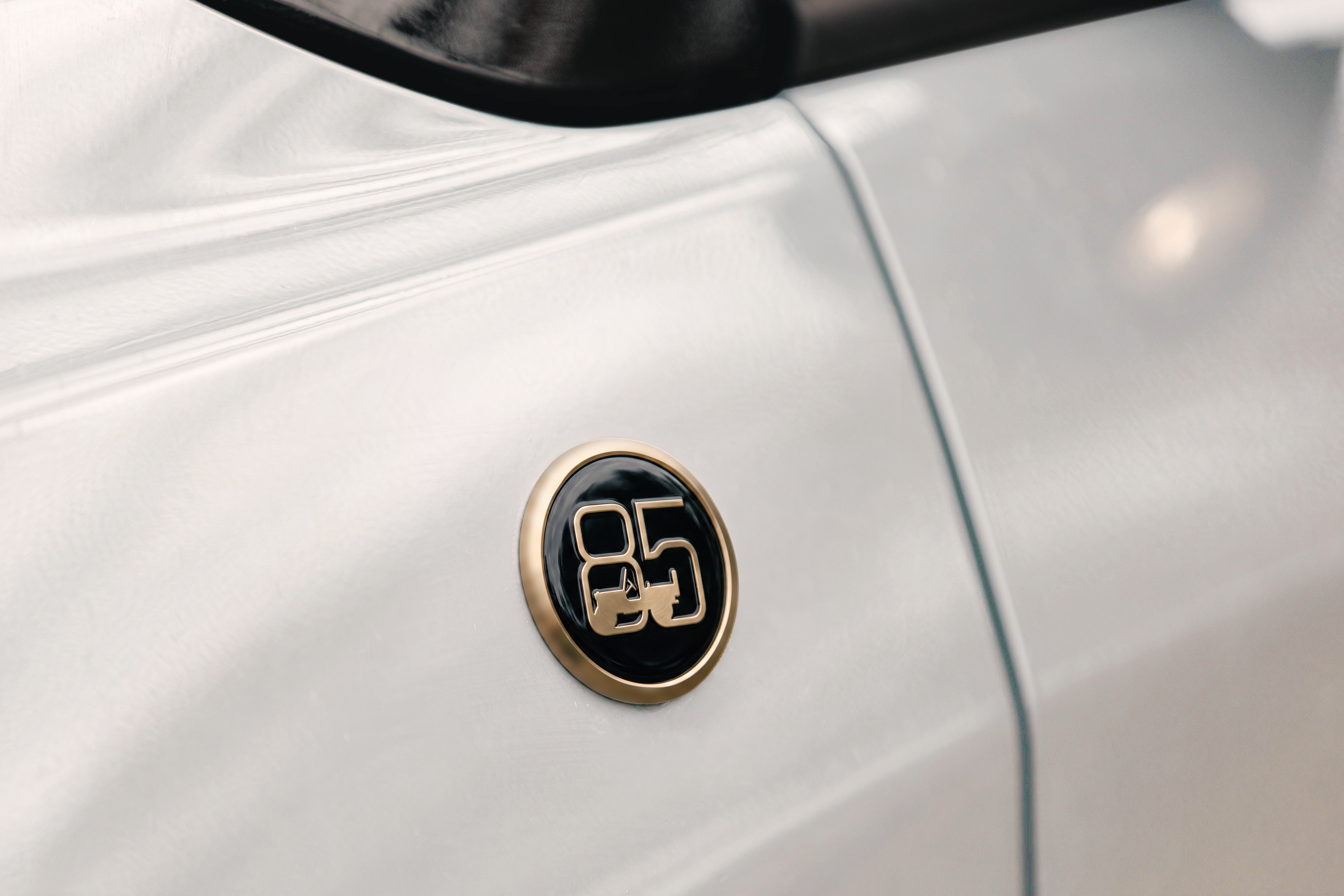 Emblema oval dourado e preto com o número 85 estilizado, fixado na lateral de um carro branco perolado