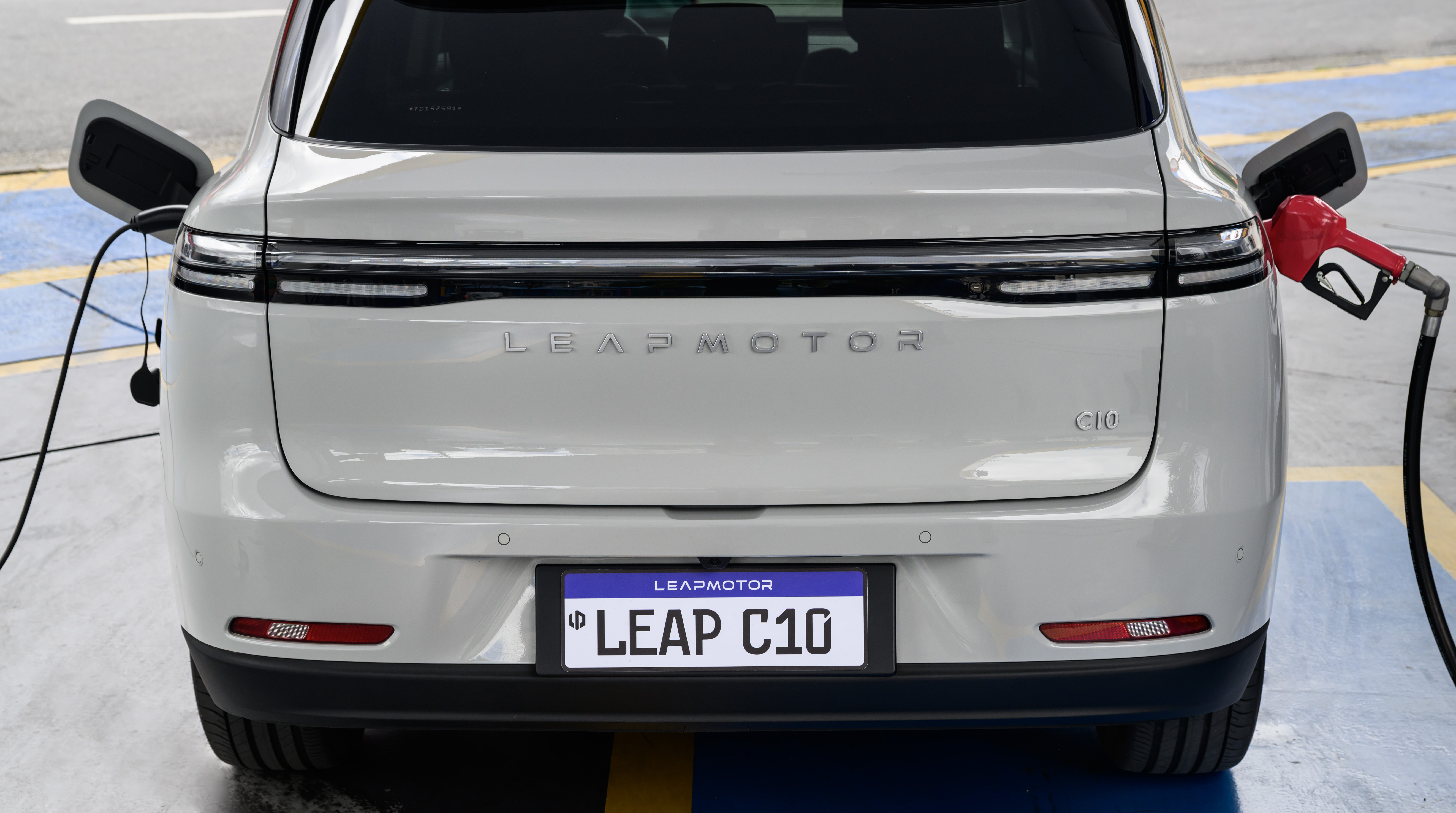 Traseira de um carro branco Leapmotor C10, com um cabo de carregamento elétrico conectado à esquerda e um bico de bomba de combustível vermelho inserido à direita, simbolizando a transição energética. A placa exibe LEAP C10