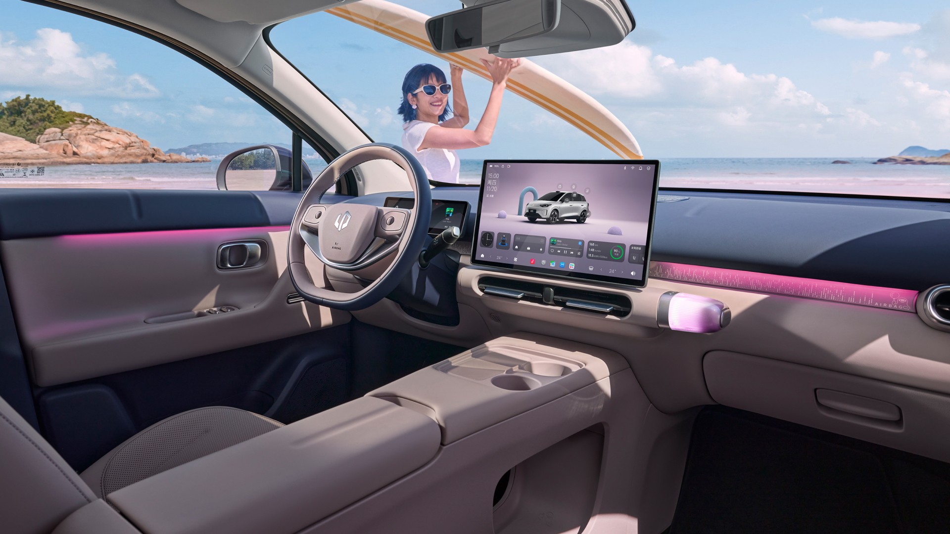 Interior de um carro moderno com painel bege e iluminação ambiente rosa. Uma mulher sorridente com óculos de sol e camiseta branca segura uma prancha de surfe no teto, com uma praia rochosa ao fundo. A tela central do carro exibe um SUV branco