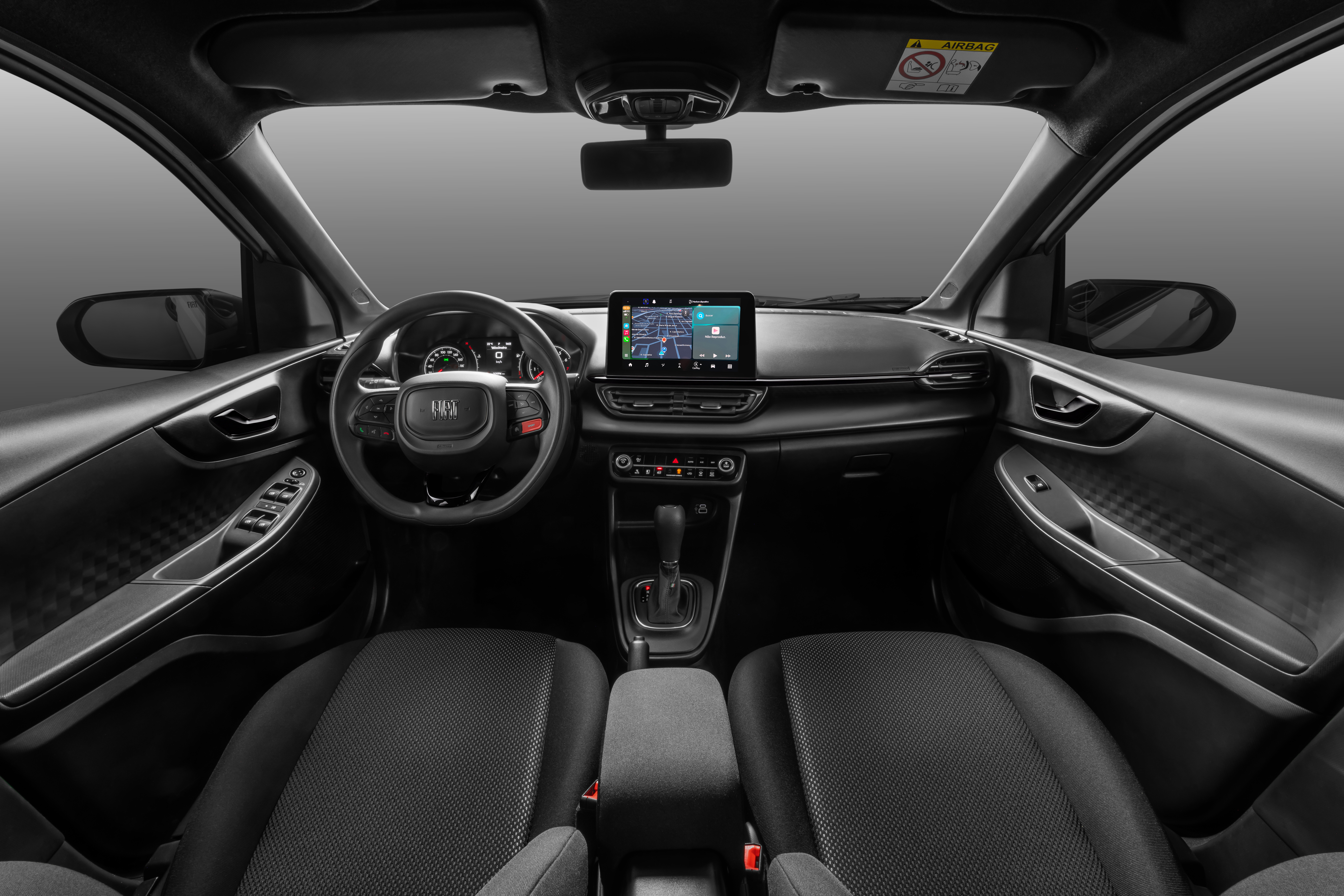 Interior de um carro moderno, com bancos pretos de tecido, volante multifuncional e painel escuro. Uma tela multimídia central exibe um mapa de navegação.
