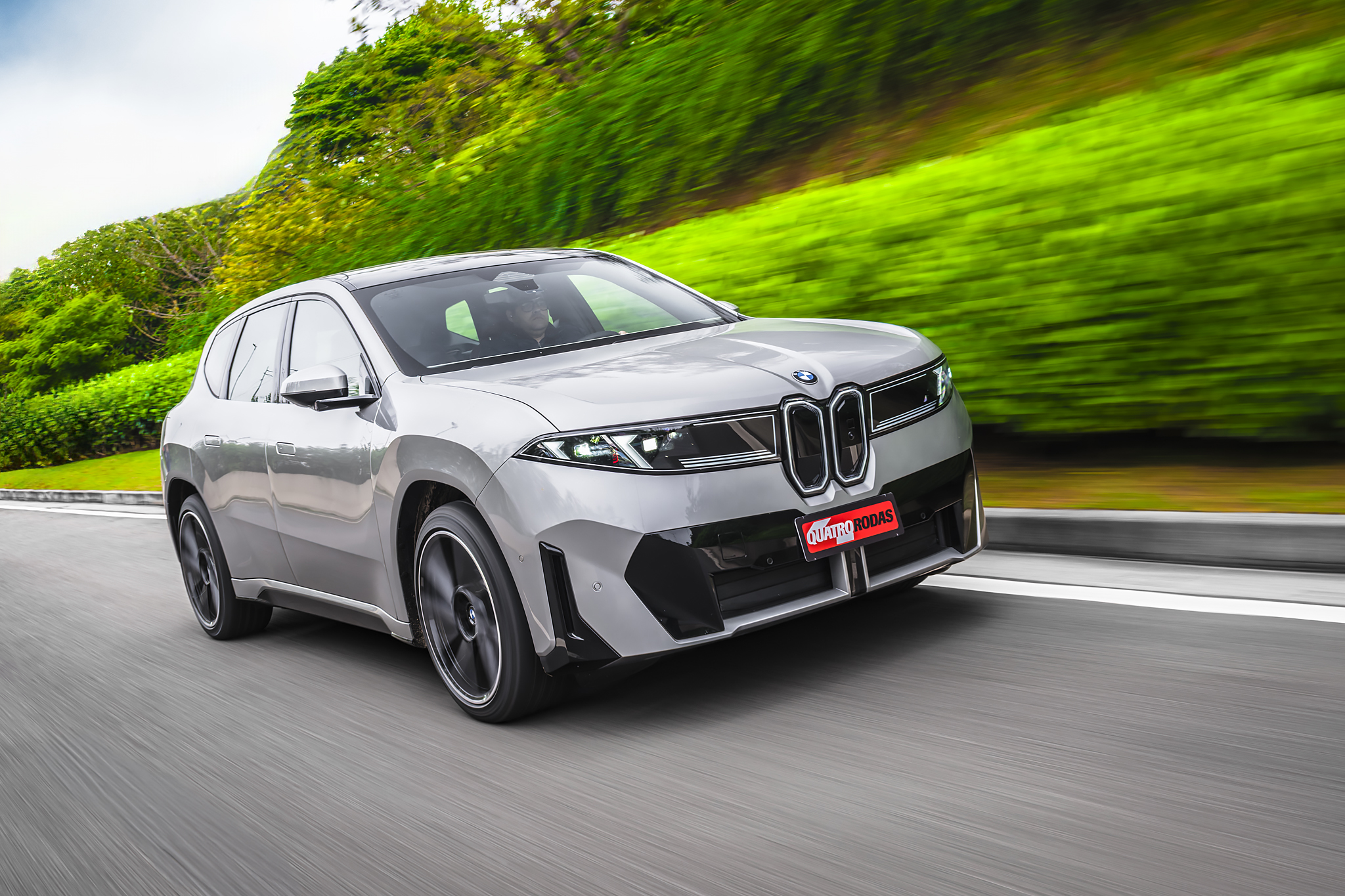 SUV BMW iX cinza claro em movimento rápido numa estrada, com vegetação verde borrada ao fundo e céu claro