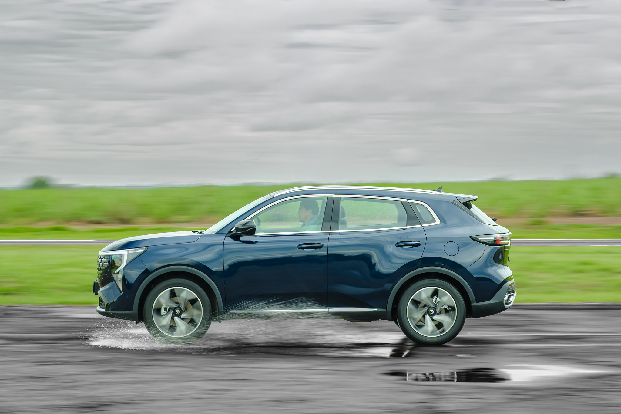 SUV azul escuro em movimento, passando por uma poça dágua na estrada, com respingos visíveis. O fundo desfocado mostra um campo verde e céu nublado