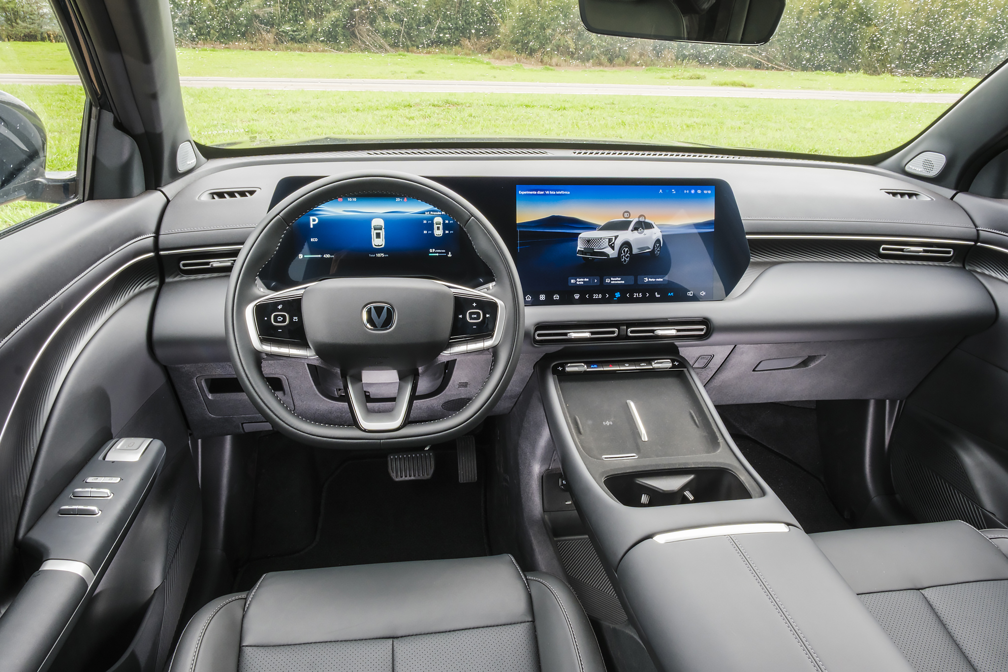 Interior moderno de um carro, com volante multifuncional e dois grandes monitores digitais. O monitor central exibe um SUV branco em uma estrada, enquanto o do painel mostra informações do veículo. Bancos de couro escuro e detalhes prateados complementam o design elegante.