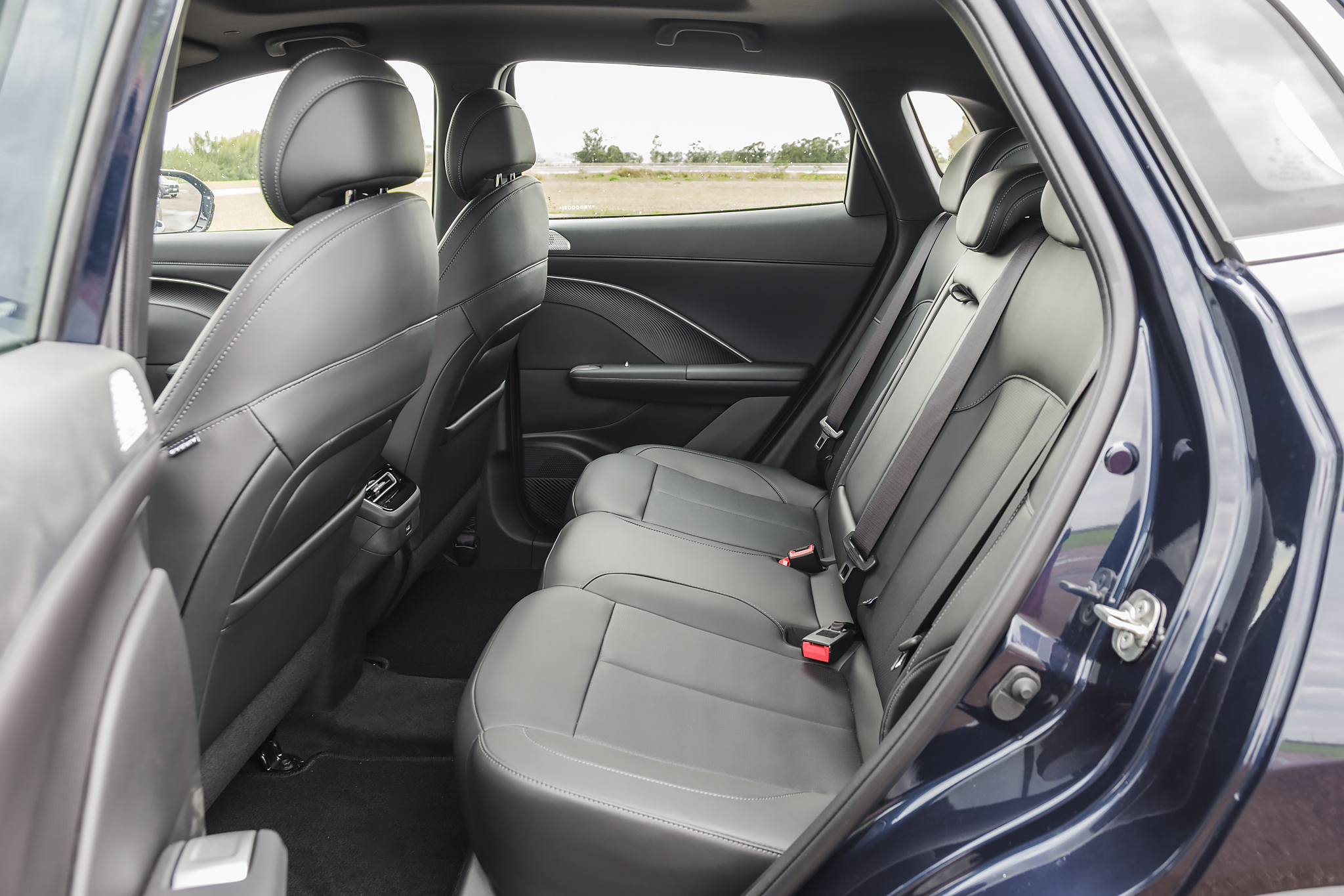 Interior de um carro azul escuro, mostrando os bancos traseiros de couro preto com cintos de segurança e fivelas vermelhas. O banco do passageiro dianteiro também é visível, com um bolso na parte de trás. A janela traseira exibe uma paisagem embaçada com vegetação e céu claro. A porta traseira está aberta, revelando a dobradiça e o mecanismo de travamento. O carpete do chão é preto.