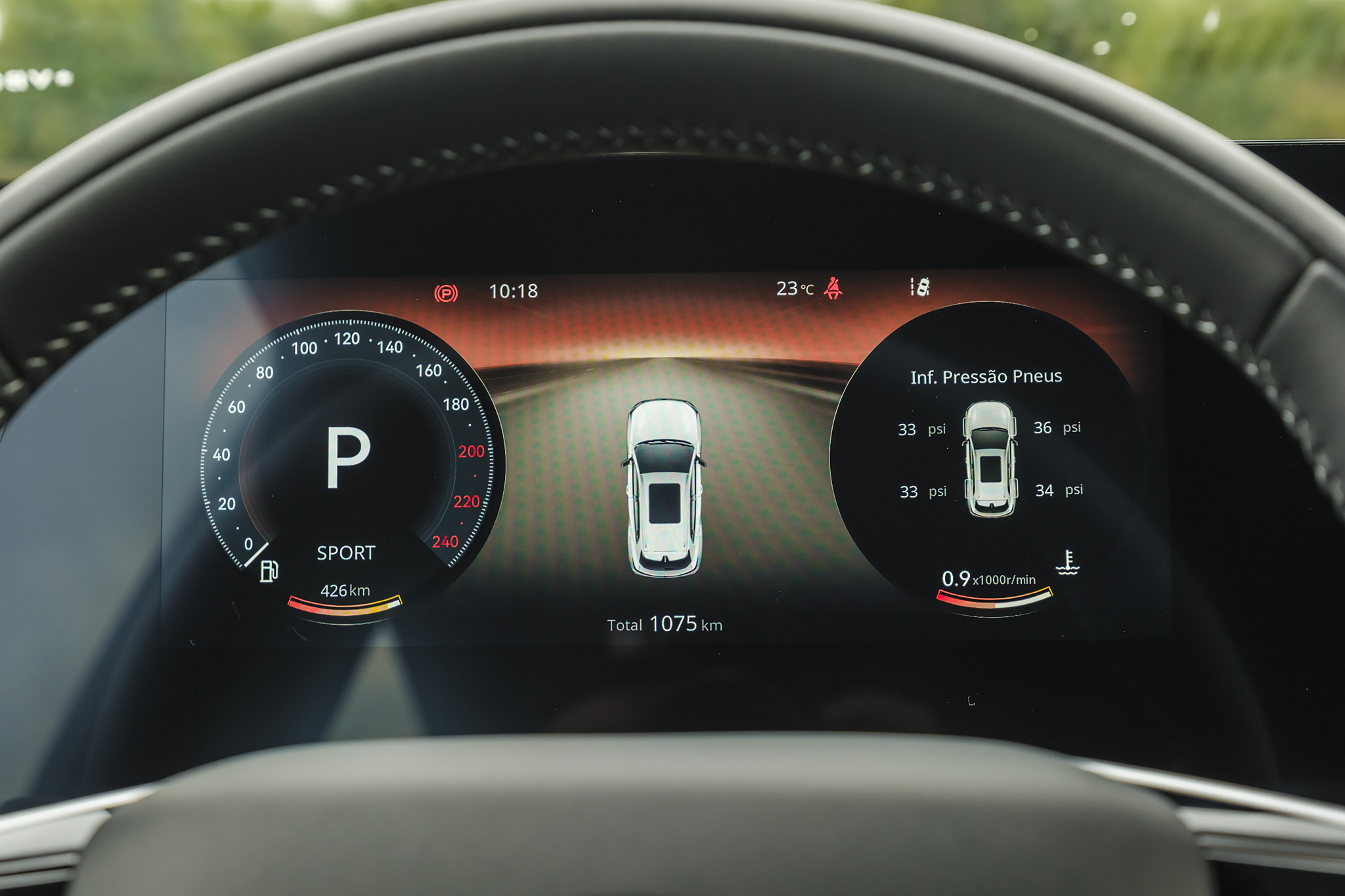 Painel digital de carro com velocímetro à esquerda marcando P e modo SPORT, autonomia de 426km. No centro, um carro branco em uma estrada e o total de 1075km. À direita, informações de pressão dos pneus (33, 36, 33, 34 psi) e temperatura. No topo, horário 10:18 e temperatura externa 23°C