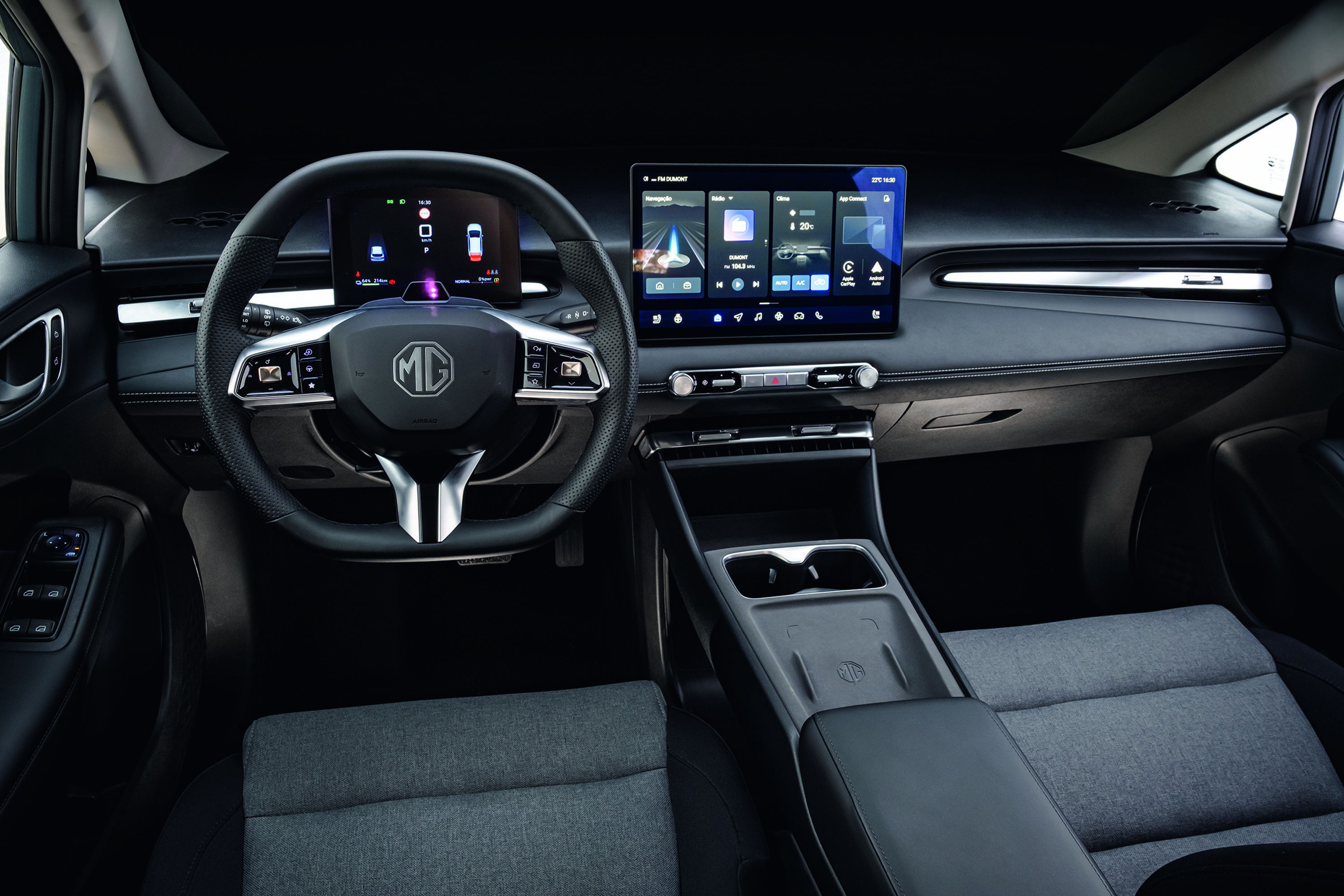 Interior de um carro moderno, com volante preto e o logotipo MG no centro. À direita, uma tela multimídia exibe um mapa de navegação e ícones de aplicativos. O painel é escuro, com detalhes prateados. Os bancos são cinza-escuro, e o console central possui porta-copos. A iluminação interna é suave
