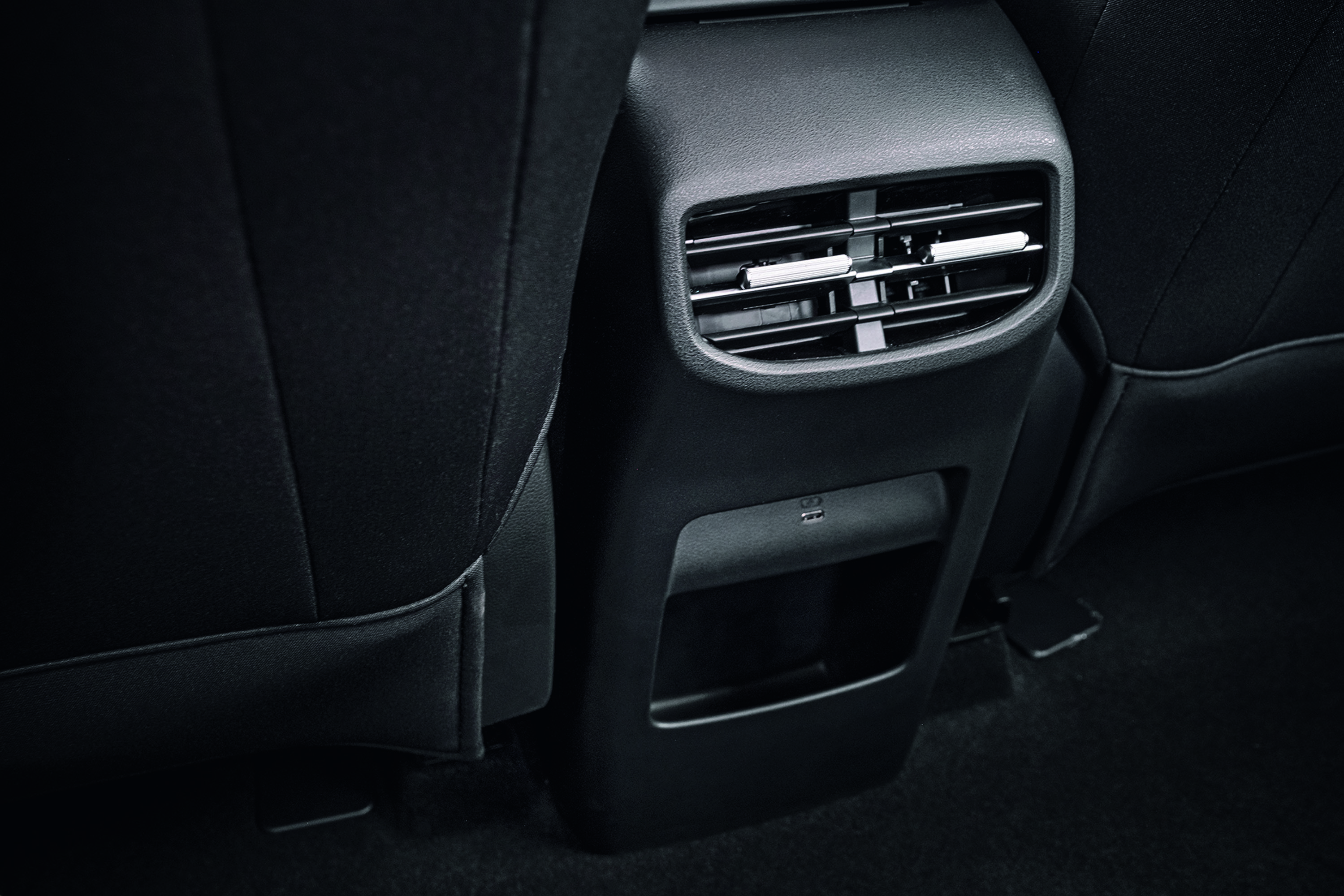 Parte traseira de um banco de carro preto, com saídas de ar horizontais, uma porta USB-C e um pequeno porta-objetos abaixo