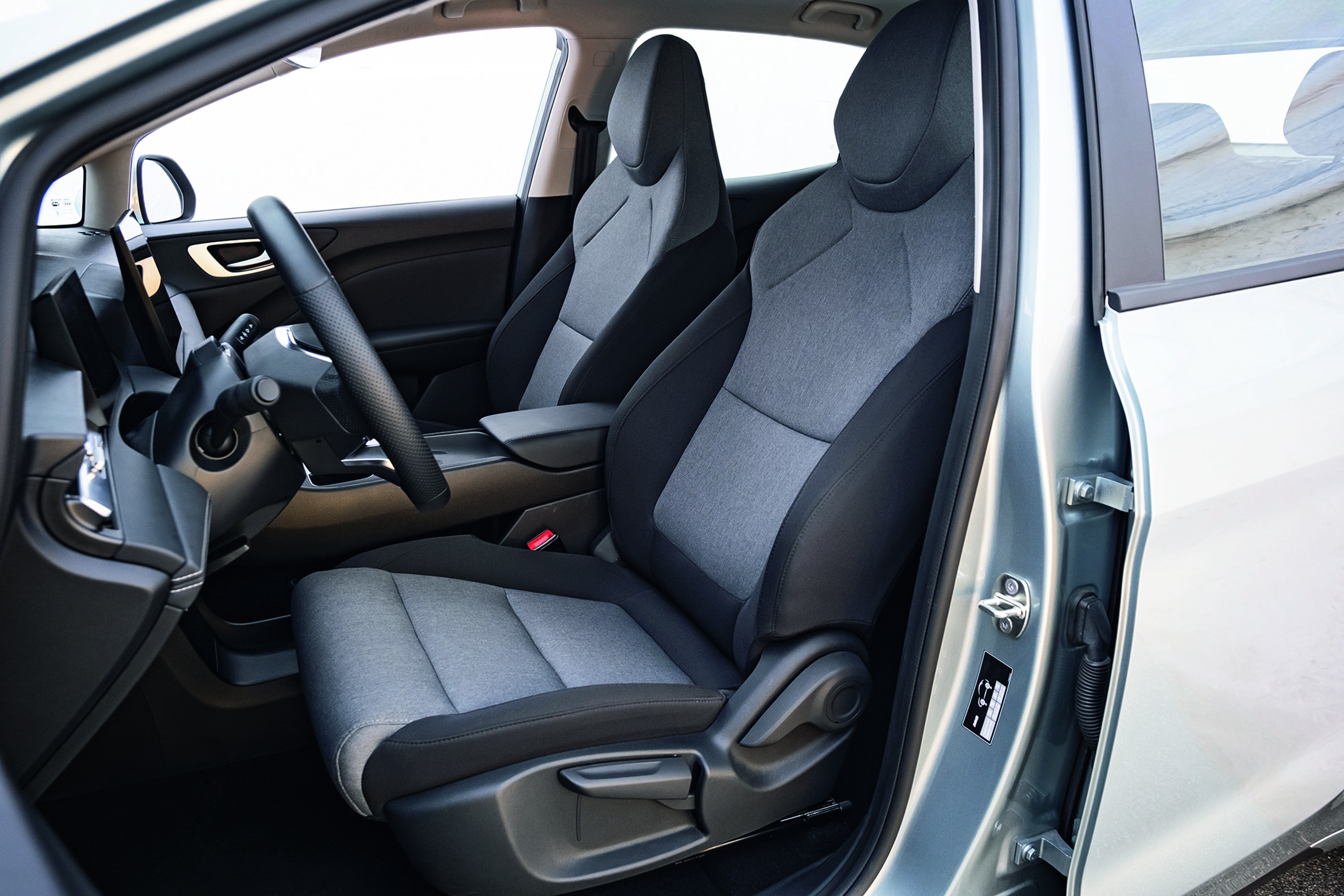 Interior de um carro com a porta do motorista aberta, revelando o volante preto e os bancos dianteiros em tecido cinza claro e escuro. O banco do passageiro também é visível, com o mesmo padrão. O ambiente é claro, com luz natural entrando pelas janelas.