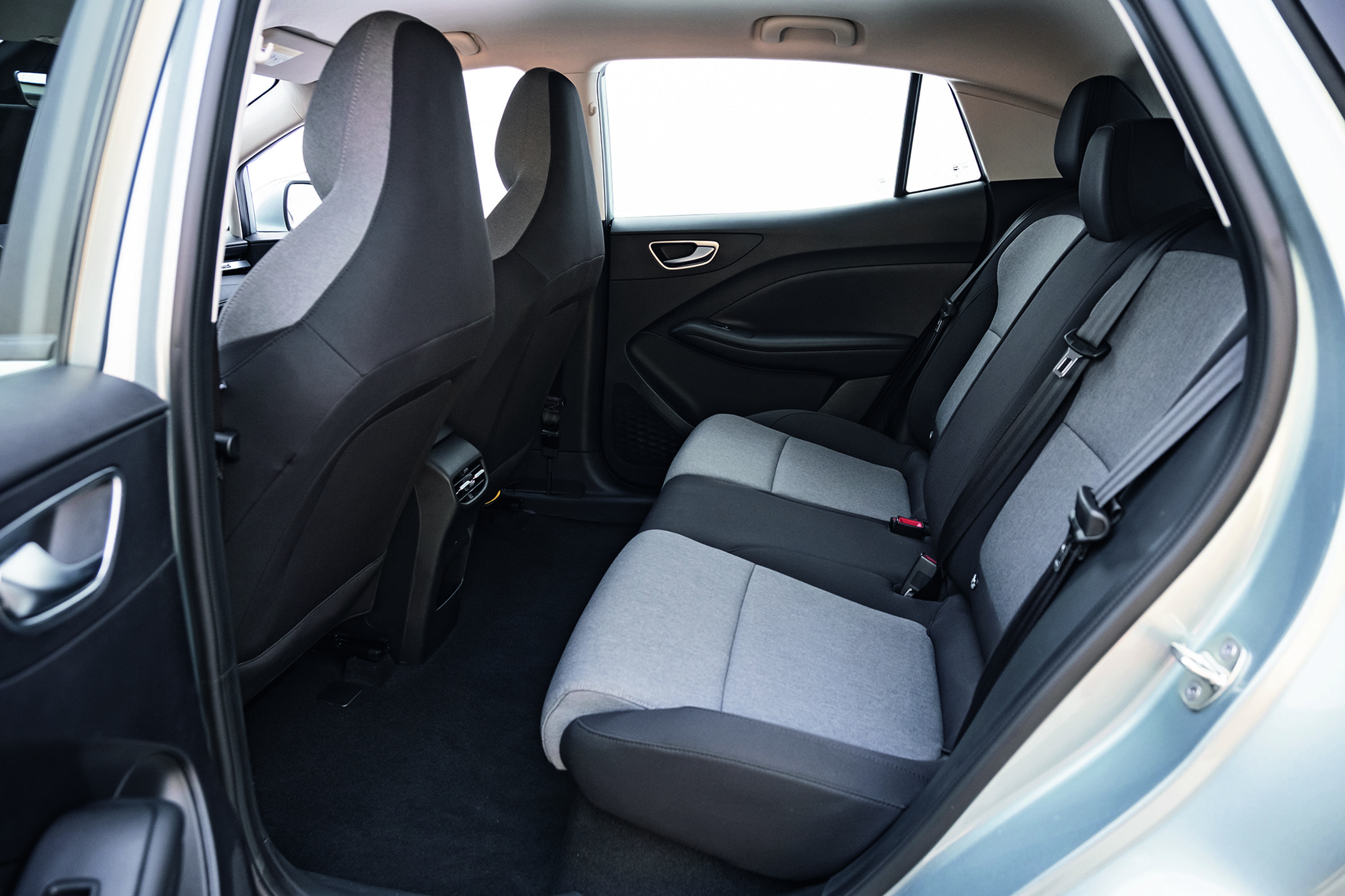 Interior de um carro com bancos traseiros em tecido cinza claro e escuro, cintos de segurança visíveis. Os bancos dianteiros, também em tecido escuro, aparecem parcialmente. O piso é preto e as portas têm detalhes em cinza escuro e puxadores cromados. A janela traseira mostra um fundo branco