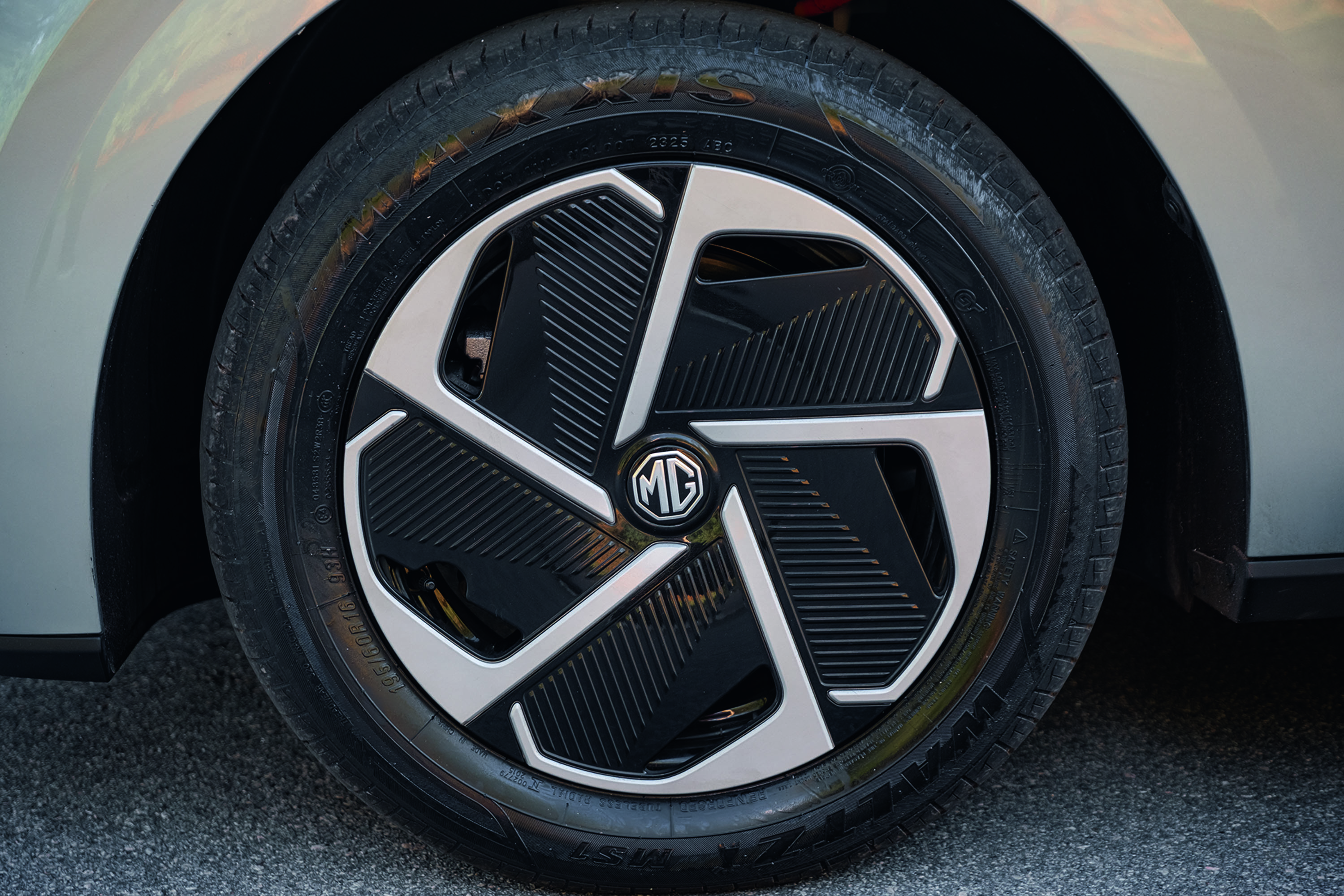 Roda de carro com pneu preto e calota prateada e preta, com o logotipo MG no centro. O carro é cinza claro e está sobre asfalto escuro