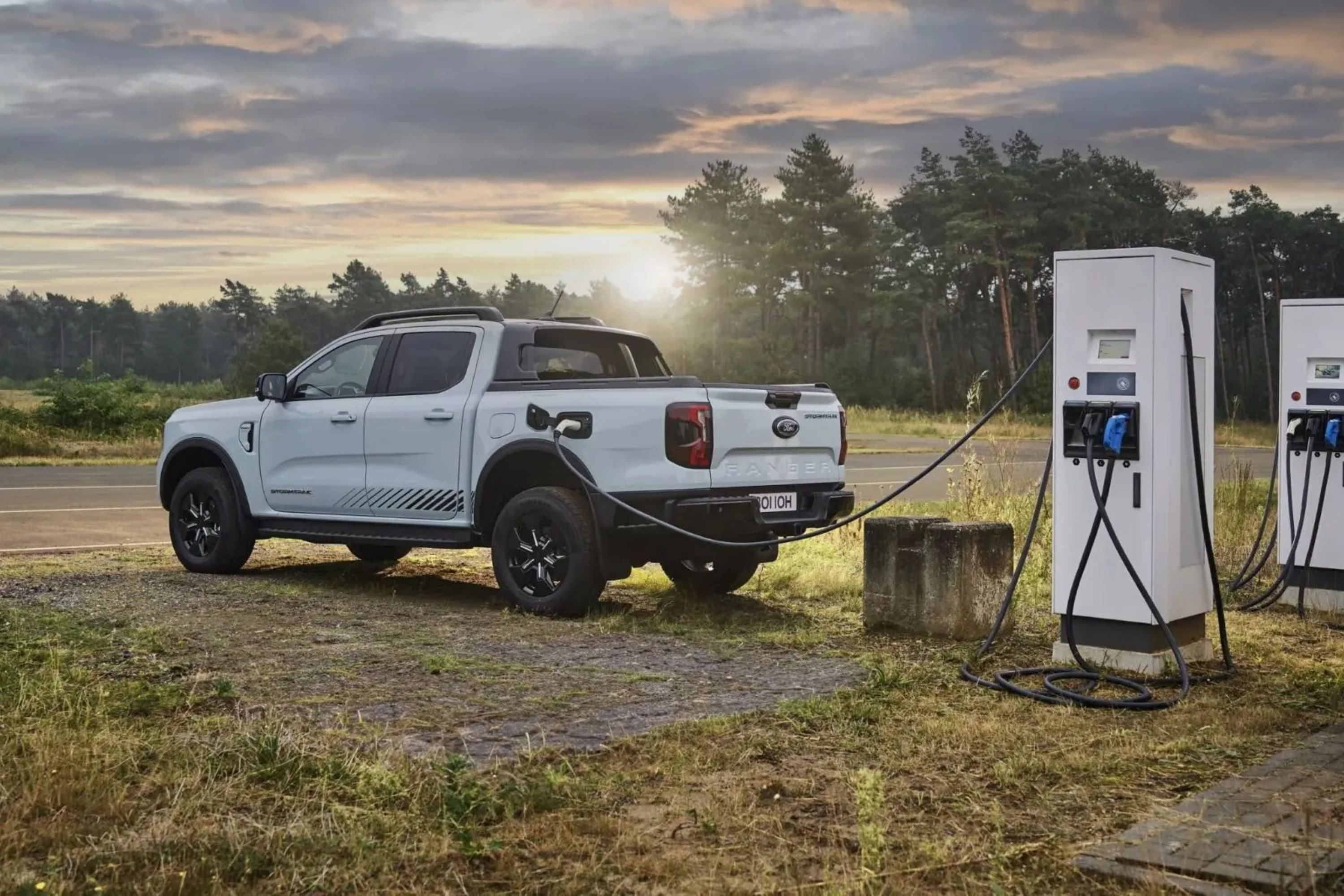 Picape branca Ford Ranger estacionada em área de terra e grama, conectada a um carregador elétrico branco. O sol se põe no horizonte, com árvores ao fundo e um céu nublado