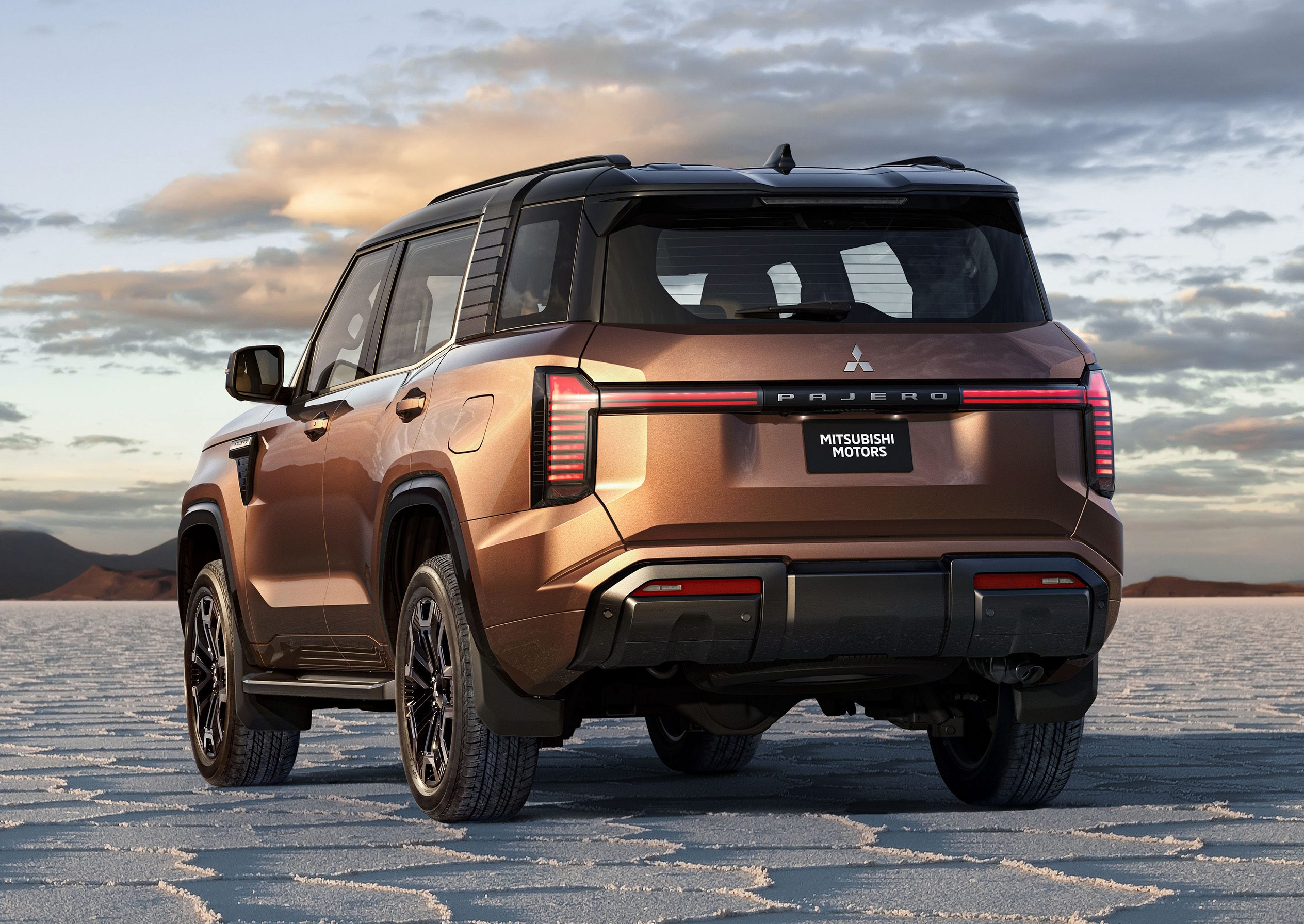 SUV Mitsubishi Pajero Sport marrom-cobre com teto preto, visto por trás, parado em um deserto de sal com padrões hexagonais no chão. Montanhas escuras ao fundo e céu azul com nuvens esparsas ao entardecer