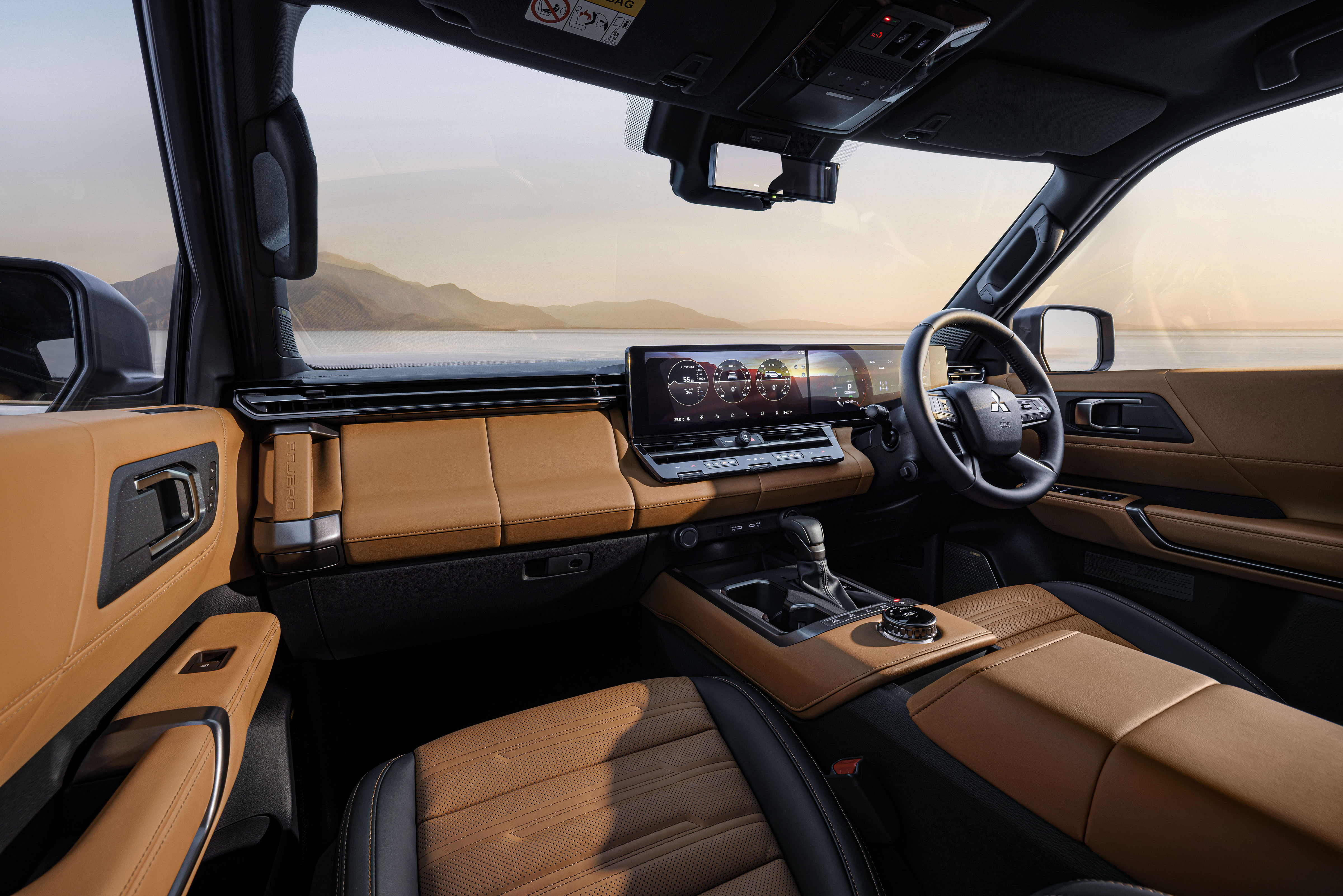 Interior de um carro moderno com estofamento marrom-claro e detalhes pretos. O painel possui uma tela digital ampla e o volante é multifuncional. Ao fundo, uma paisagem montanhosa sob um céu claro ao entardecer