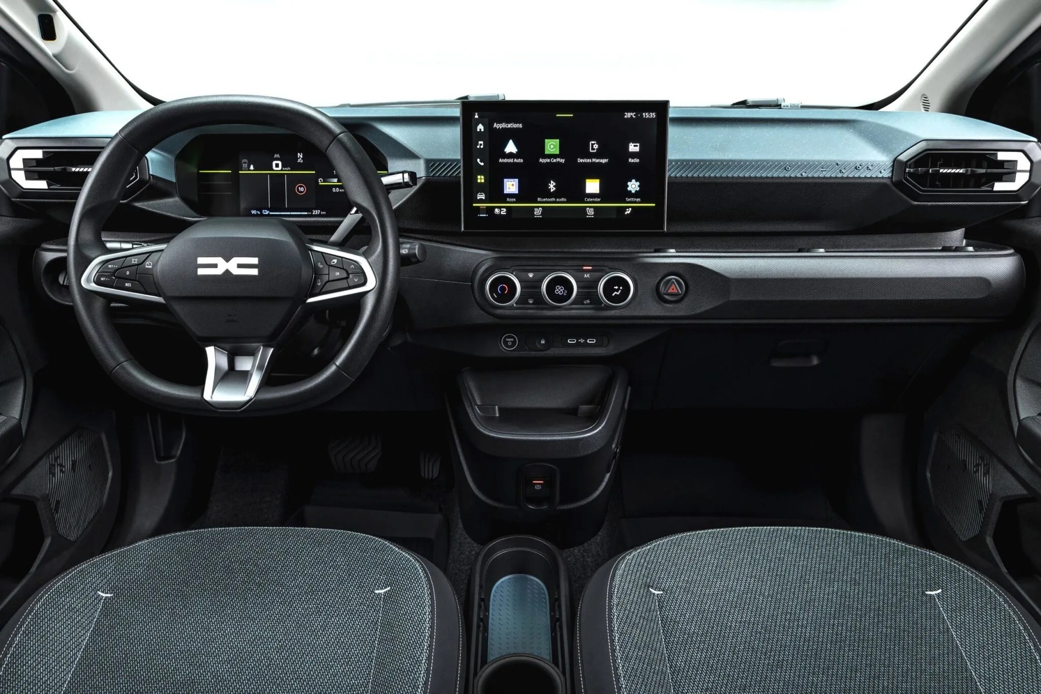 Interior de um carro Dacia moderno, com volante preto e logotipo Dacia, painel digital e tela multimídia central exibindo aplicativos. O painel tem detalhes em azul claro texturizado e saídas de ar retangulares. Os bancos dianteiros são cinza escuro com costuras brancas