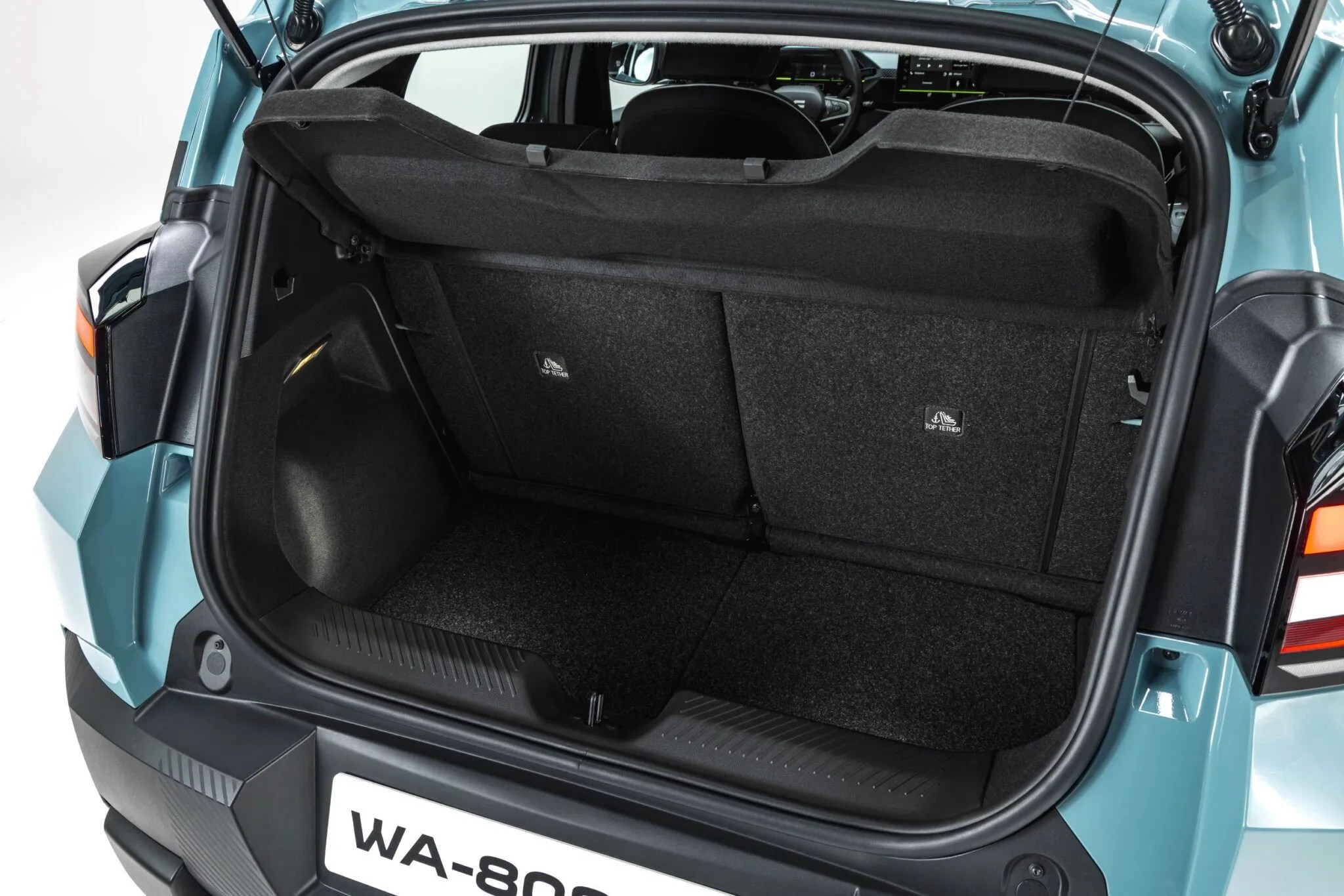Porta-malas aberto de um carro azul-esverdeado, com o assoalho e as laterais revestidos em carpete cinza escuro. O banco traseiro está rebatido, e a tampa do porta-malas está levantada, revelando o interior espaçoso e vazio do compartimento. A placa do veículo, parcialmente visível, exibe WA-80.