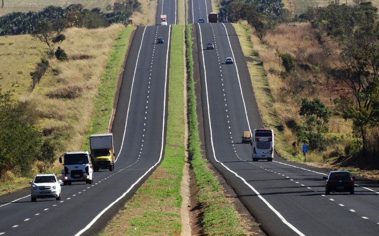 Rodovia de pista dupla com canteiro central verde, subindo em direção ao horizonte. Carros, caminhões e um ônibus trafegam em ambos os sentidos, sob céu claro e sol forte. As margens da estrada são cobertas por vegetação rasteira e algumas árvores.