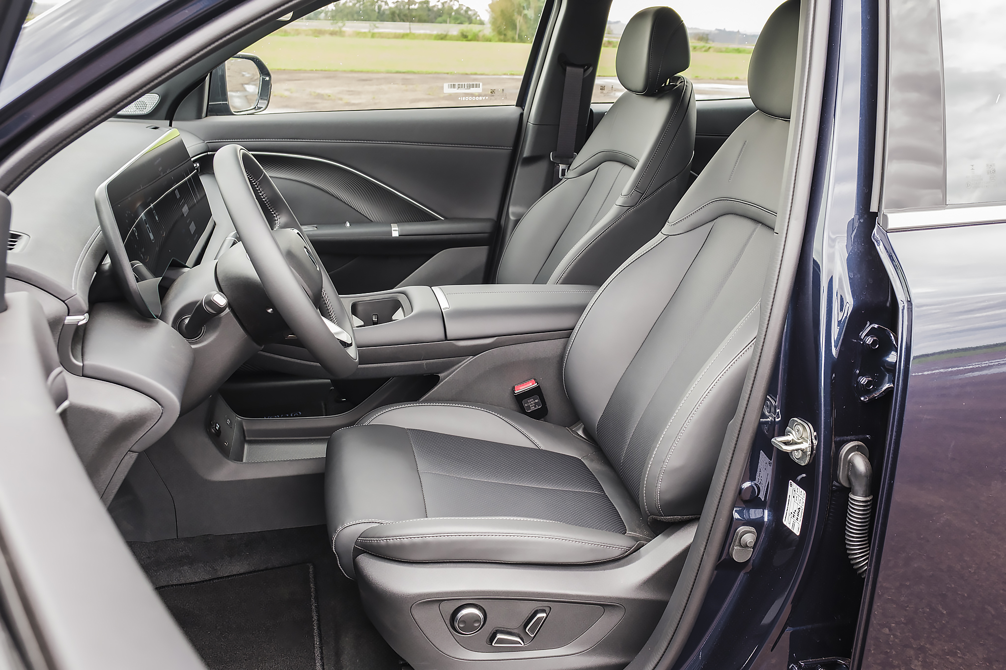 Interior de um carro azul-escuro, com a porta do motorista aberta. Volante e painel digital visíveis, bancos de couro cinza-escuro e claro, com costuras brancas. O banco do motorista possui controles elétricos na lateral. O chão é coberto por tapetes pretos. Ao fundo, um campo verde e céu claro.